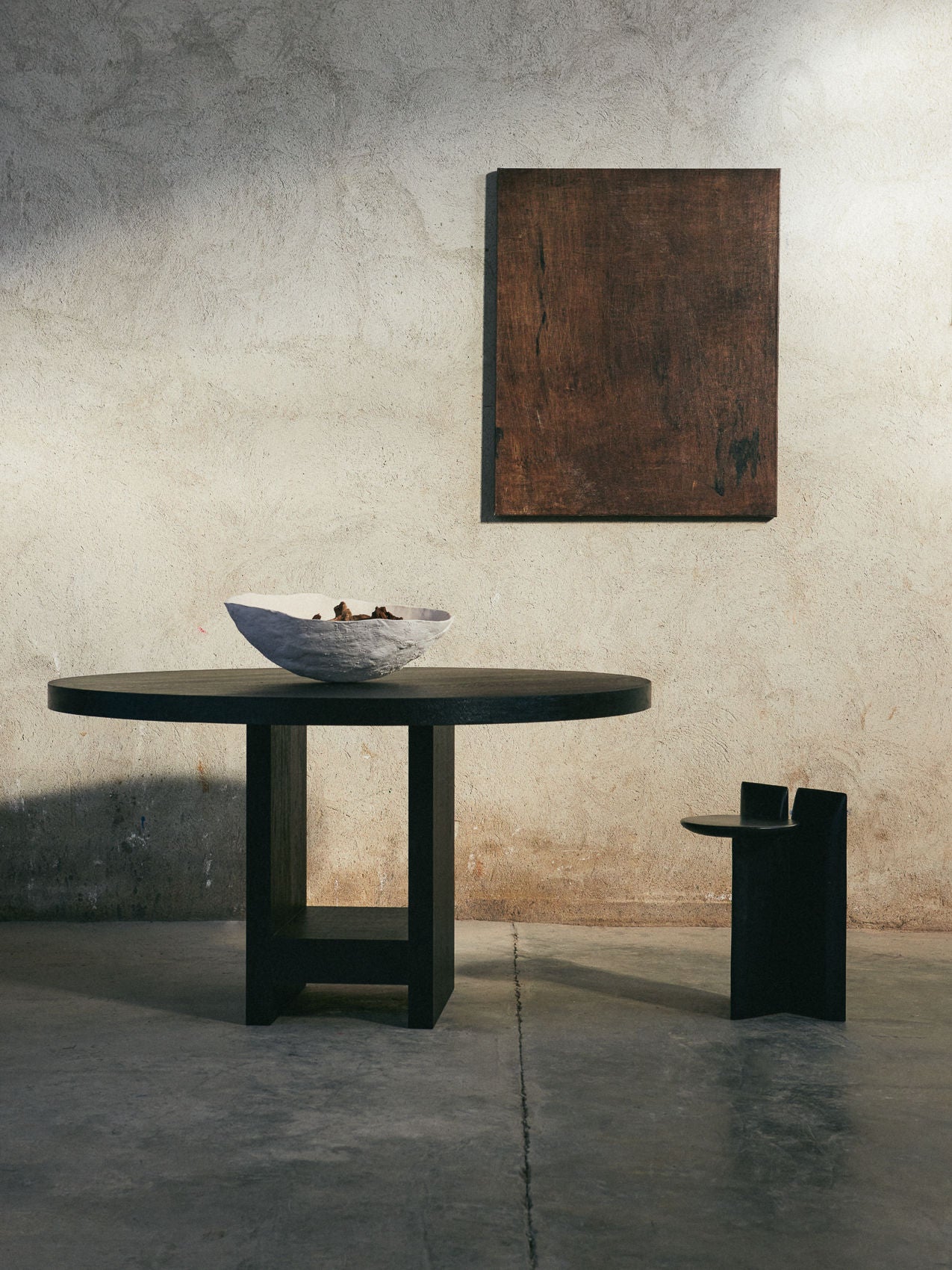 A minimalist room features the Sister Table by ASERIES OF OBSTACLES, SL, with a large light-colored bowl on top, paired with a simple wooden chair and abstract artwork on a textured beige wall. The concrete floor and soft natural light enhance the elegance of natural materials.