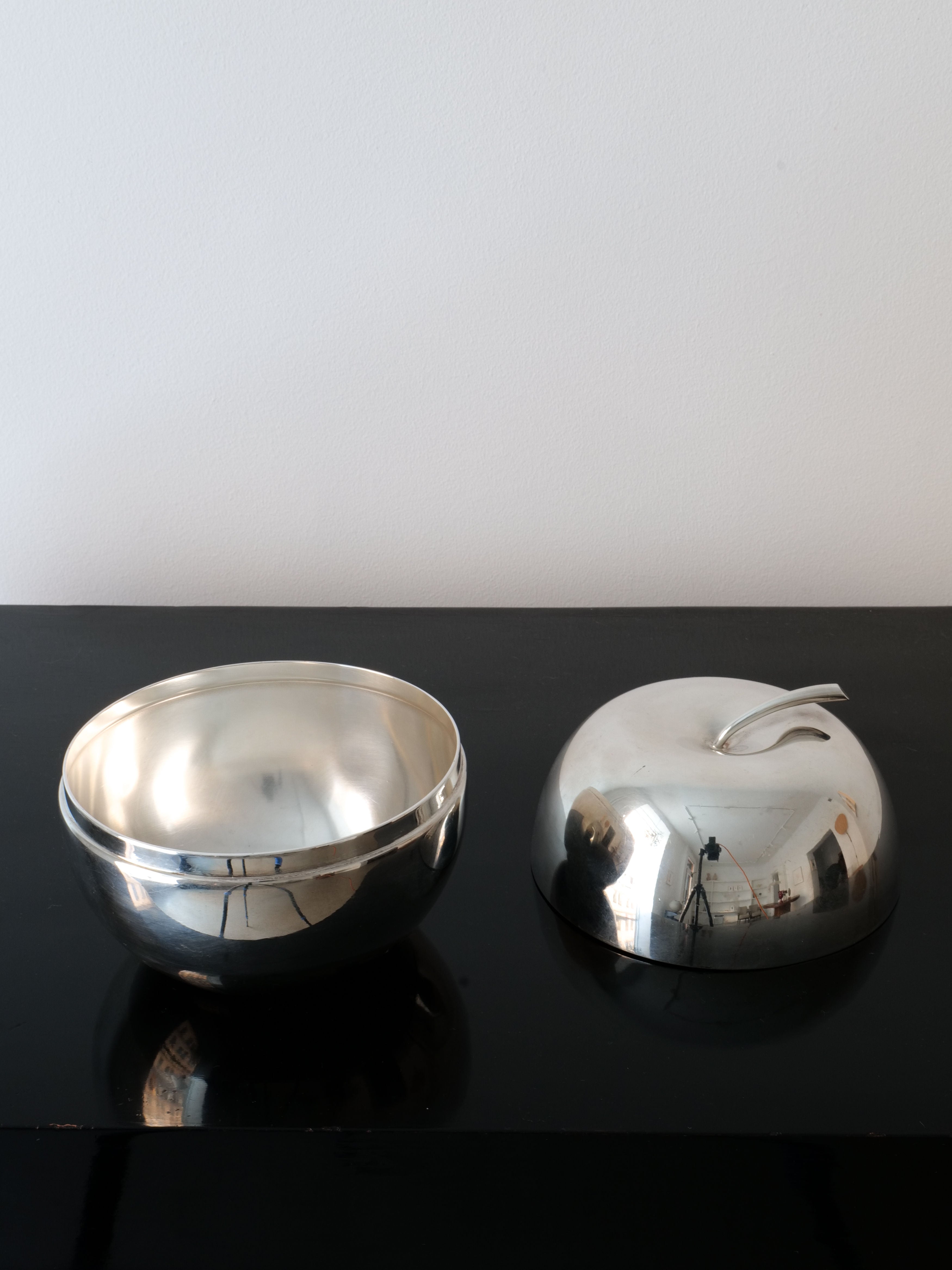 A Christofle Apple Jar by Lino Sabattini from Collection apart, featuring a polished silver bowl and its matching lid, rests on a glossy black surface. The slightly curved lid with a small handle reflects Sabattini's signature style against a plain white wall.