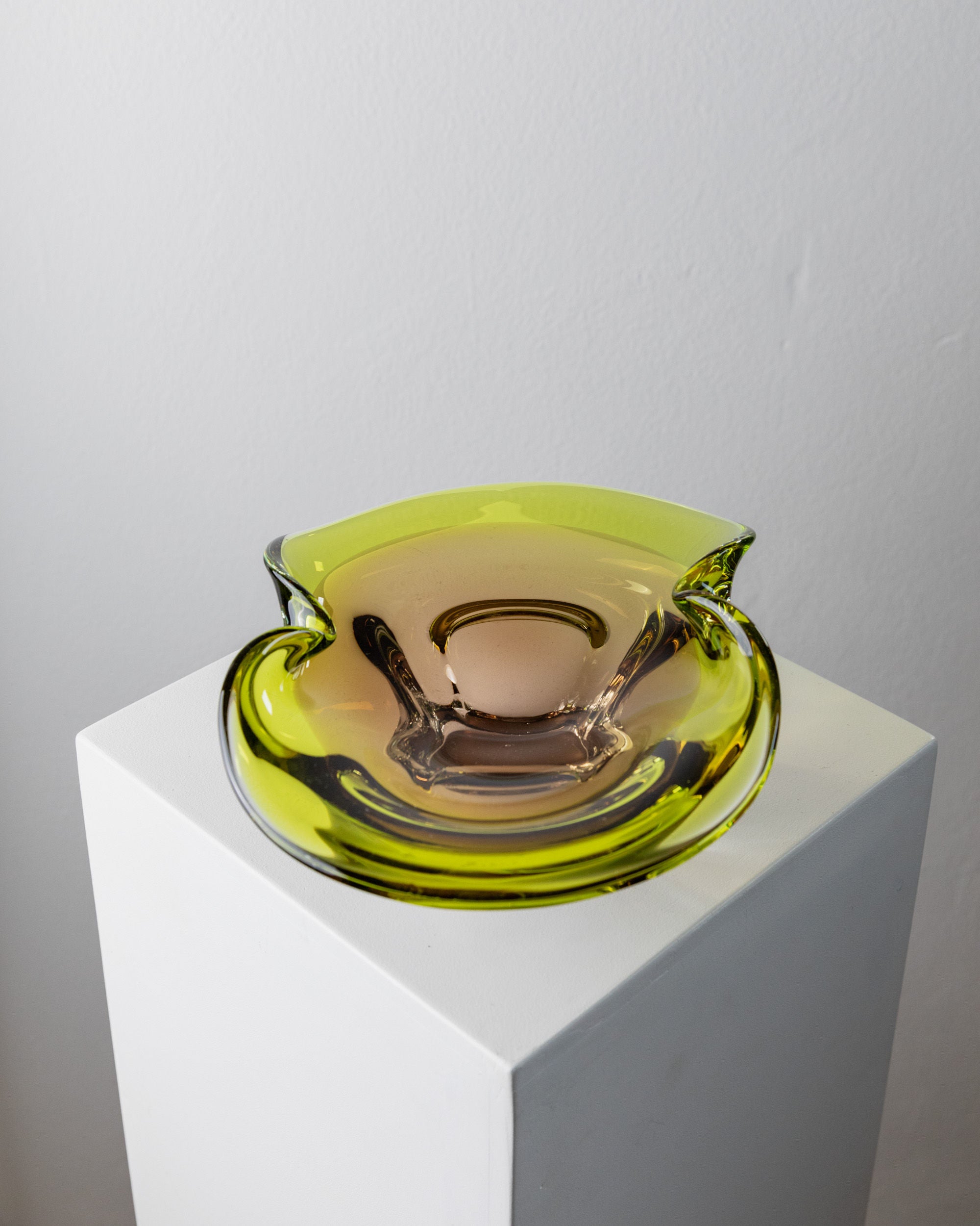 A unique Murano Glass Bowl by Bottega Jacobs from the 1970s, featuring translucent yellow-green hues and a smooth, flowing form with a central depression, sits on a white pedestal. Its abstract shape exemplifies Italian craftsmanship against a plain gray background.