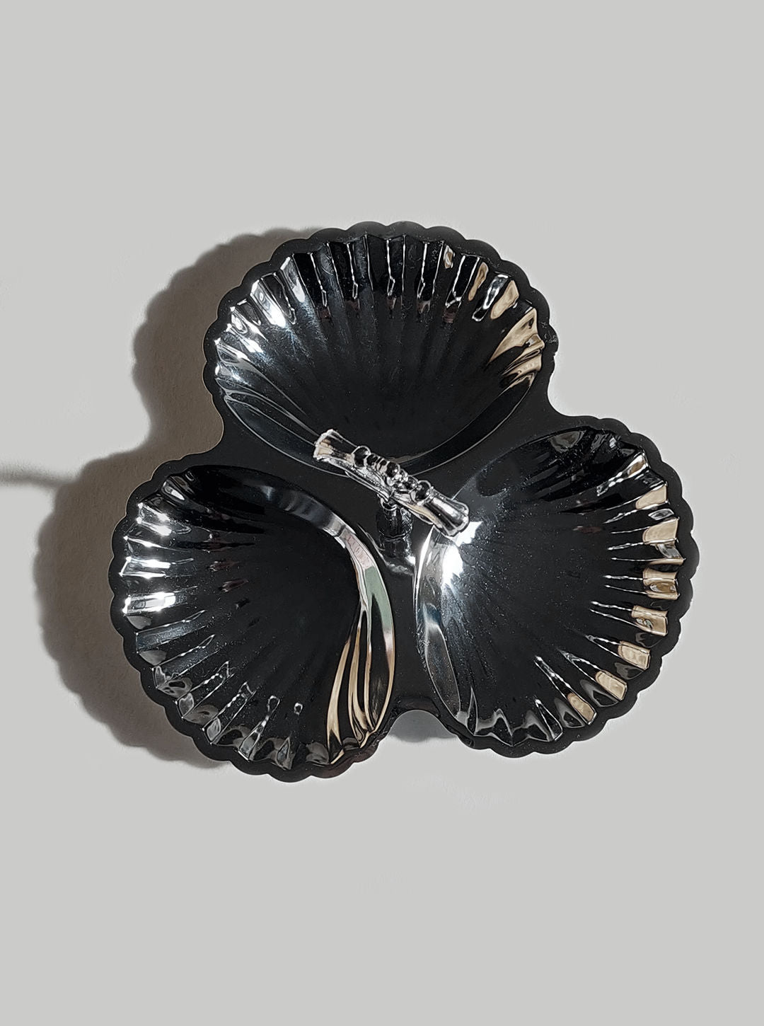 The Les Objoies Triple Shell Server, a shiny stainless steel serving dish with a sleek scalloped design, is illuminated against a neutral background. This decorative shell server features three sections and a small central handle.