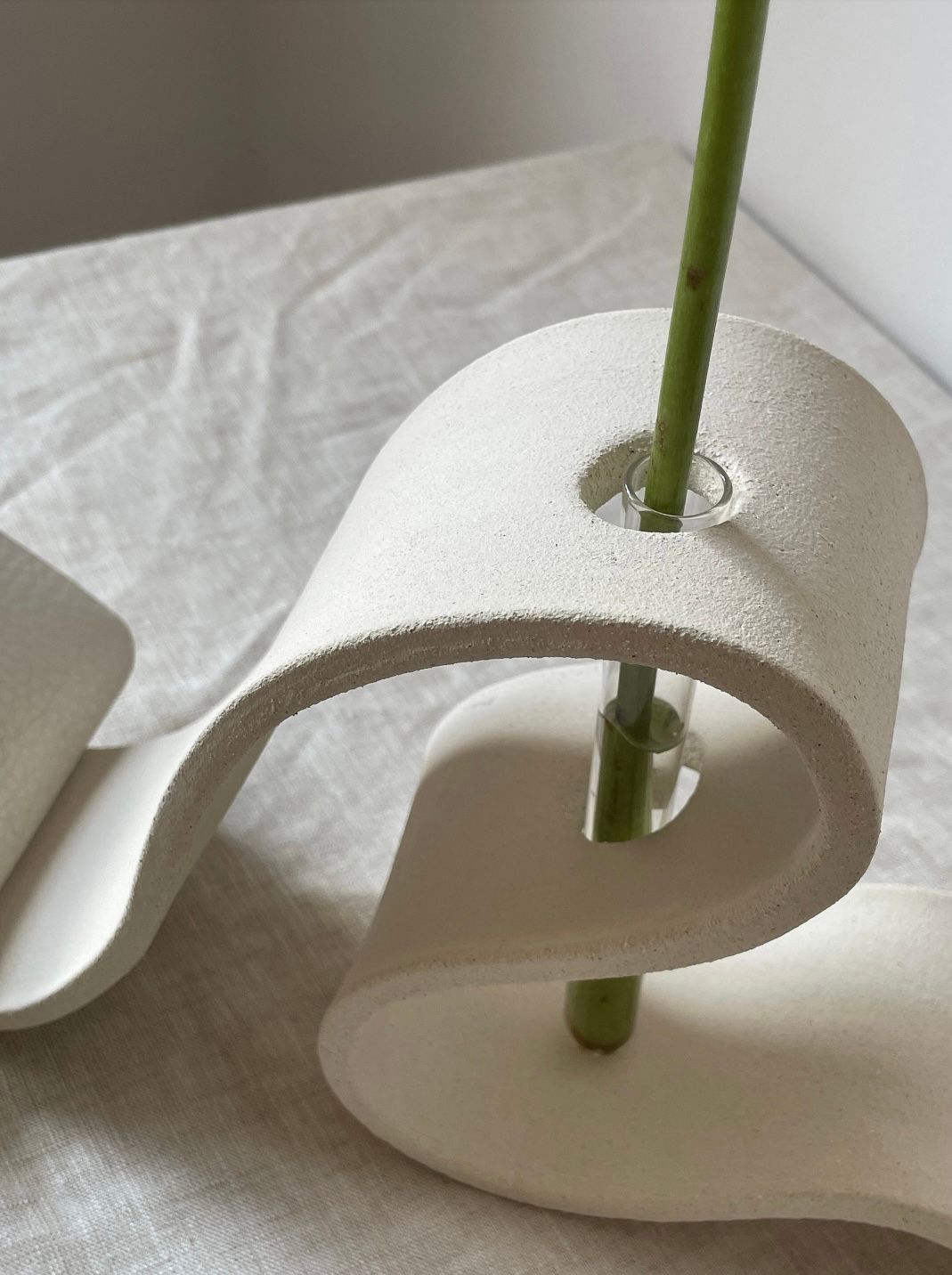 A close-up photo of a unique, modern Ribbon Vase in Off White by LESS with a wavy, white ceramic structure. A green plant stem emerges from a small glass tube inserted in the vase. The vase rests on a light-colored fabric surface with subtle creases.