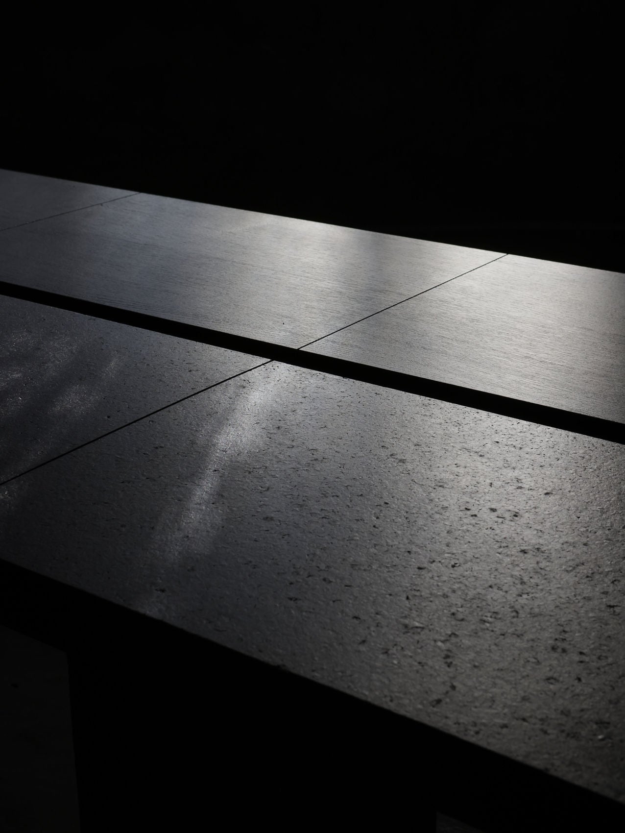 A close-up of the Obstacles Twin Oaks Dining Table showcases its dark, rectangular oak surface with a textured finish, softly illuminated by natural light. The gentle glow highlights the grain and edges against a dark background.