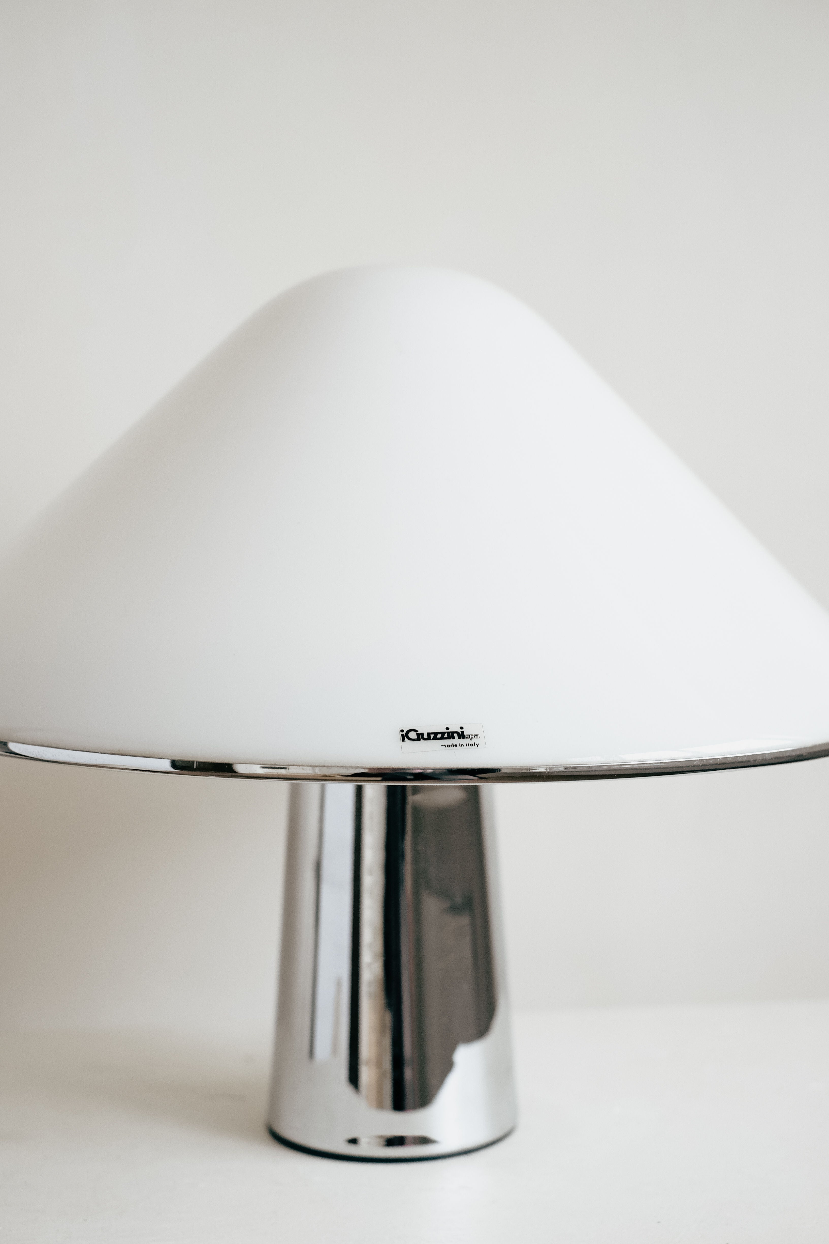 The septembre studios Harvey Guzzini Elpis Table Lamp features a shiny chrome base and wide white cone shade, inspired by the vintage classic. It sits on a white surface with a small label visible on the shade.