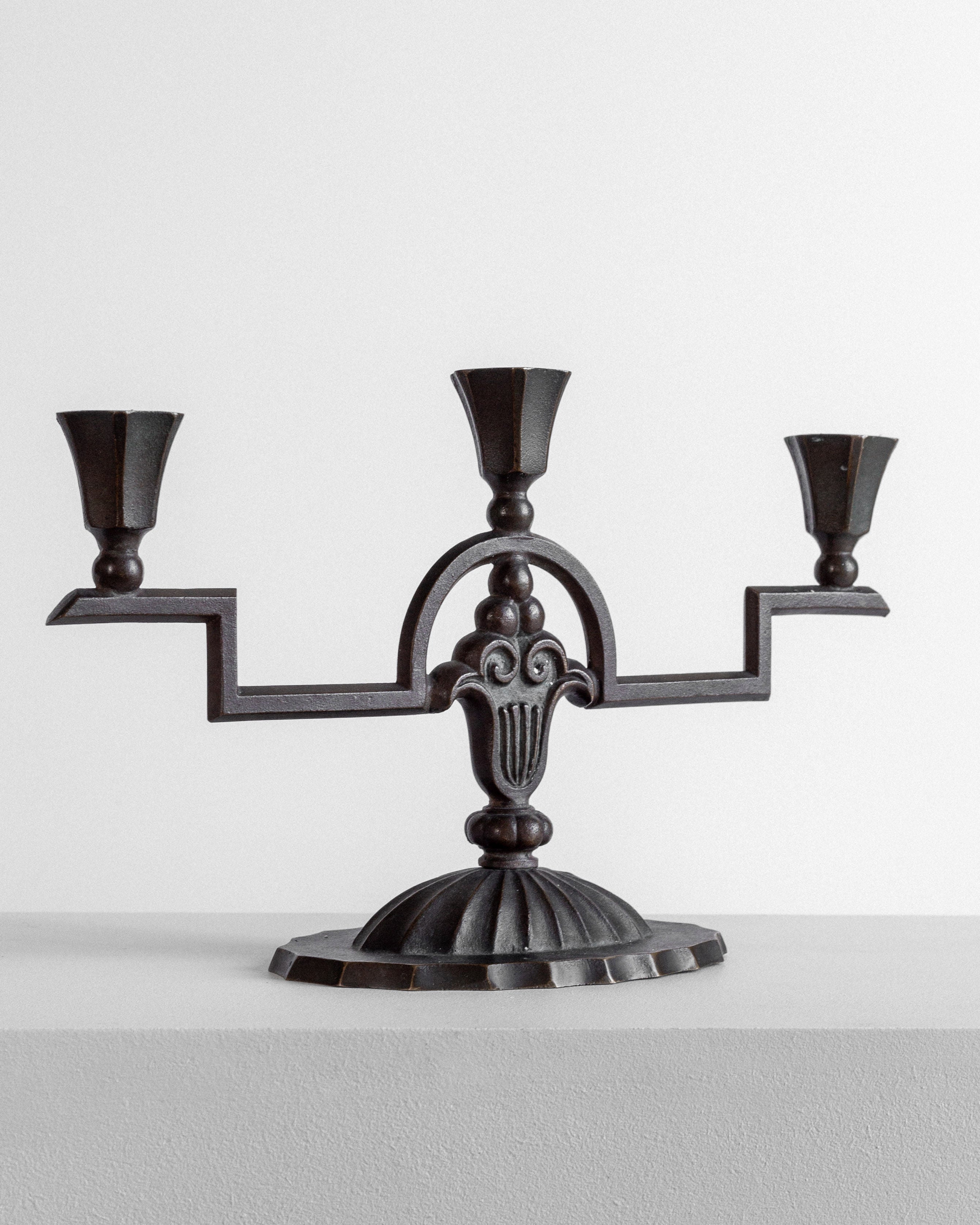 The Spigel Art Deco Bronze Candelabra from the 1930s stands on a flat surface against a plain white background. This three-armed, dark-colored piece showcases an ornate central design and a sturdy, decorative base. Each arm extends horizontally, ending in a cup to hold a candle.