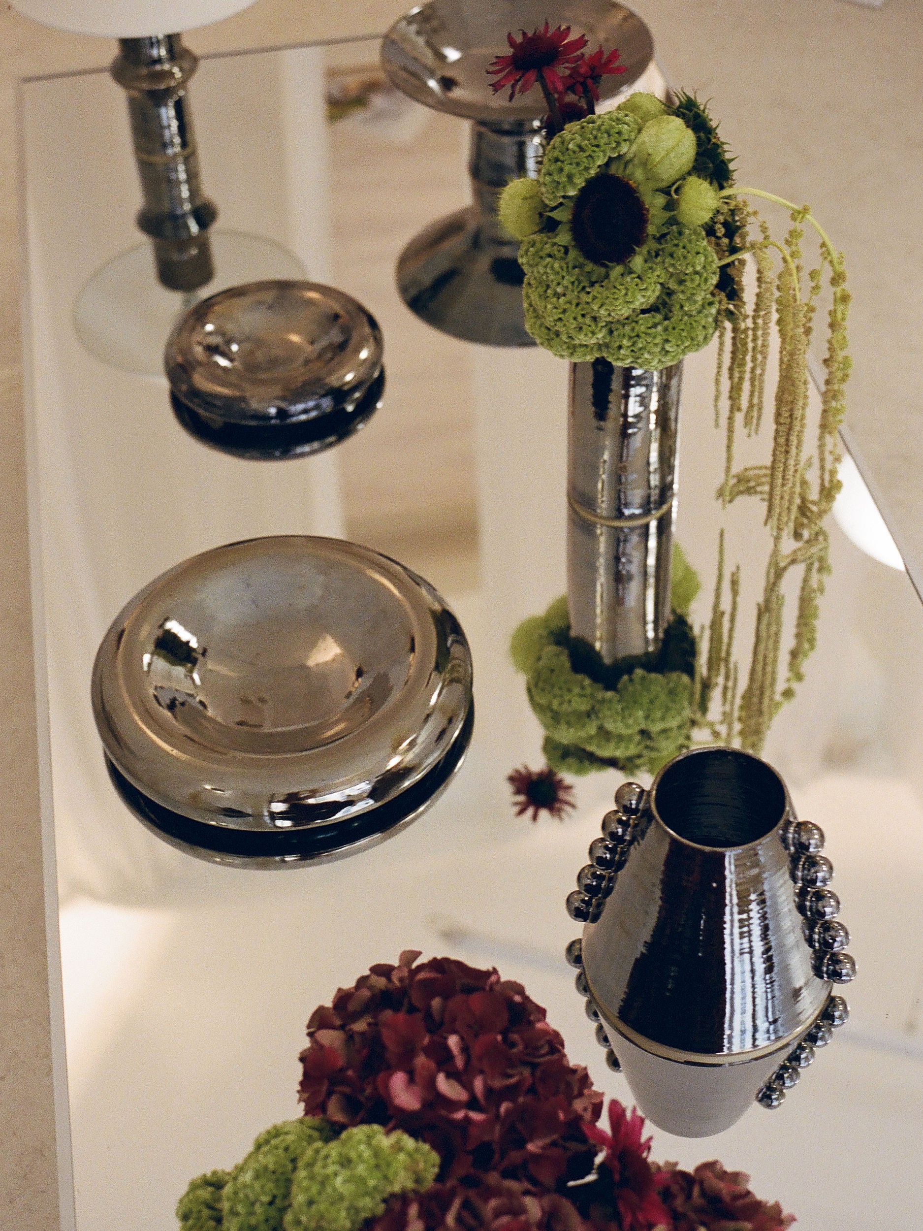 A reflective table adorned with silver metallic chrome-finished vases and bowls showcases an elegant and contemporary decor. One vase from the Landy Rakoto Ceramics collection, known as the Tubular Vase I, holds green and red flowers paired with cascading greenery, while another features a delightful arrangement of red and green blooms.