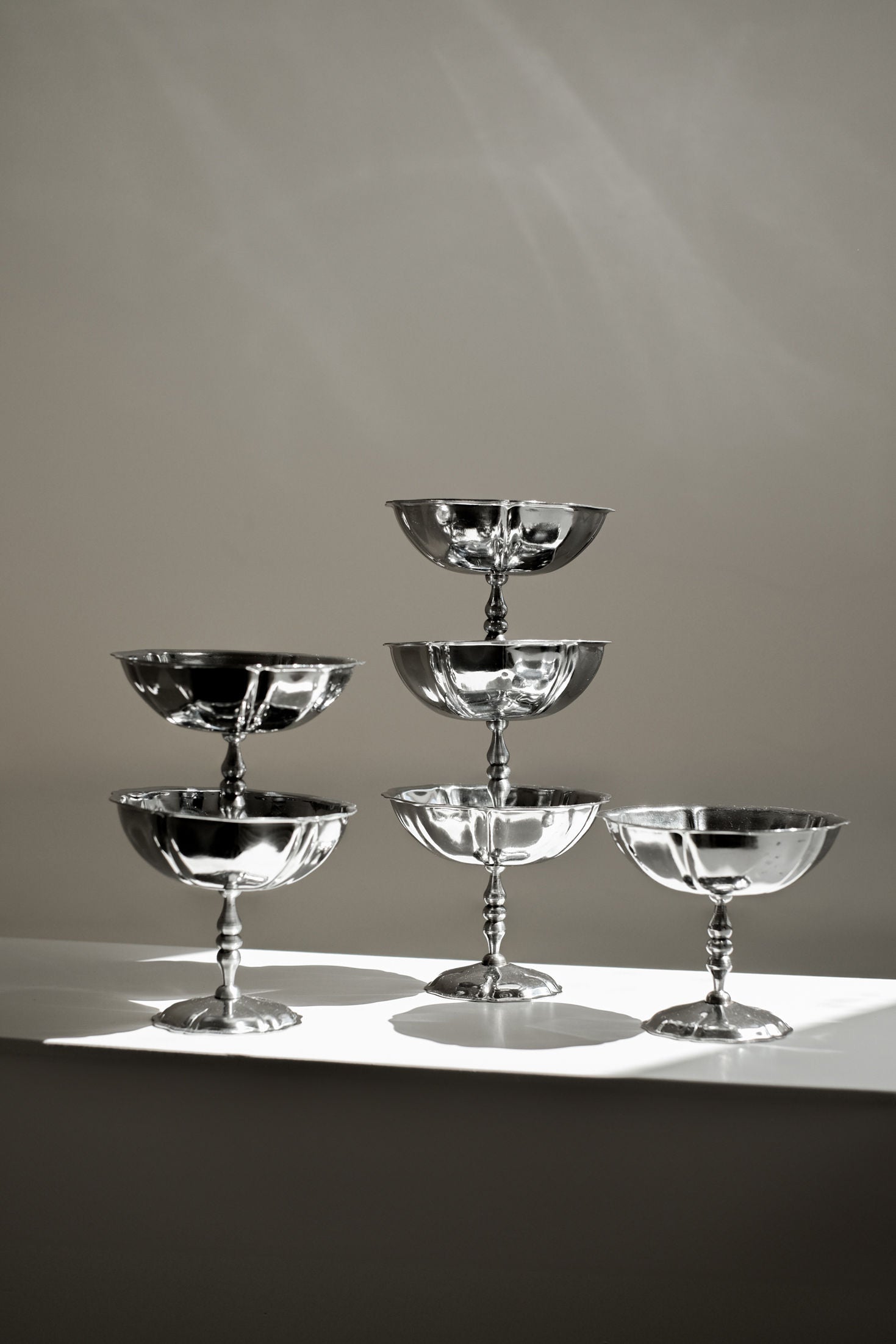 A set of six shiny stainless steel ice coupes by septembre studios is stacked in a pyramid on a white surface, with light and shadows creating reflections that enhance their vintage look.