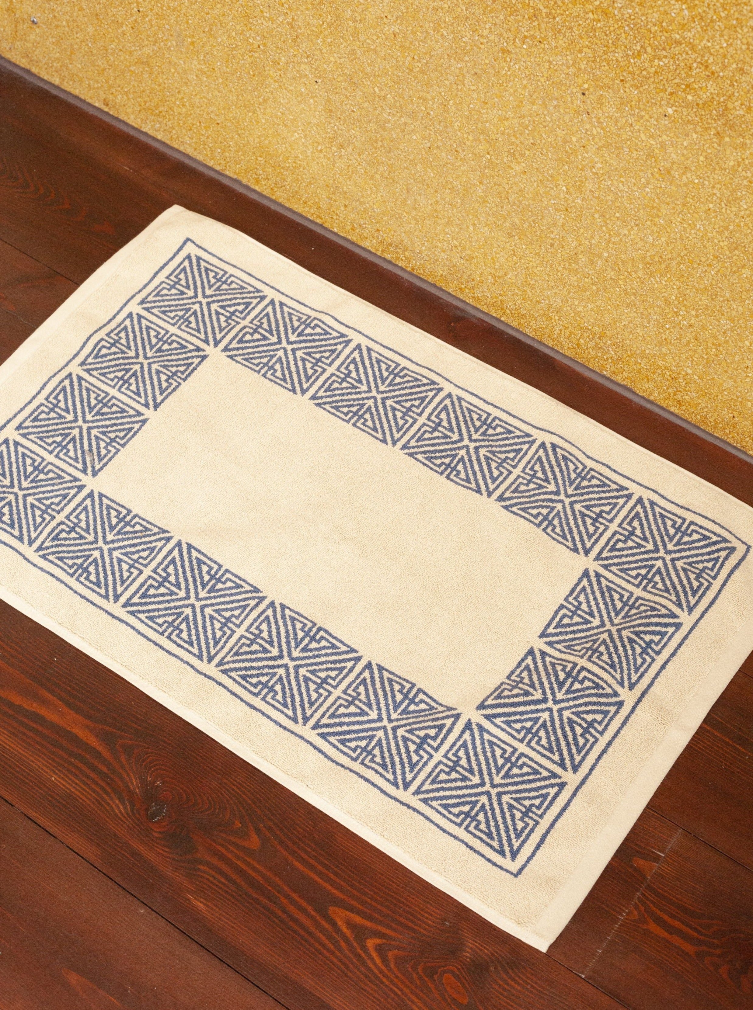 A Agnes Bath Mat by Autumn Sonata with a beige center and marine blue geometric borders, placed on a wooden floor against a textured gold wall.