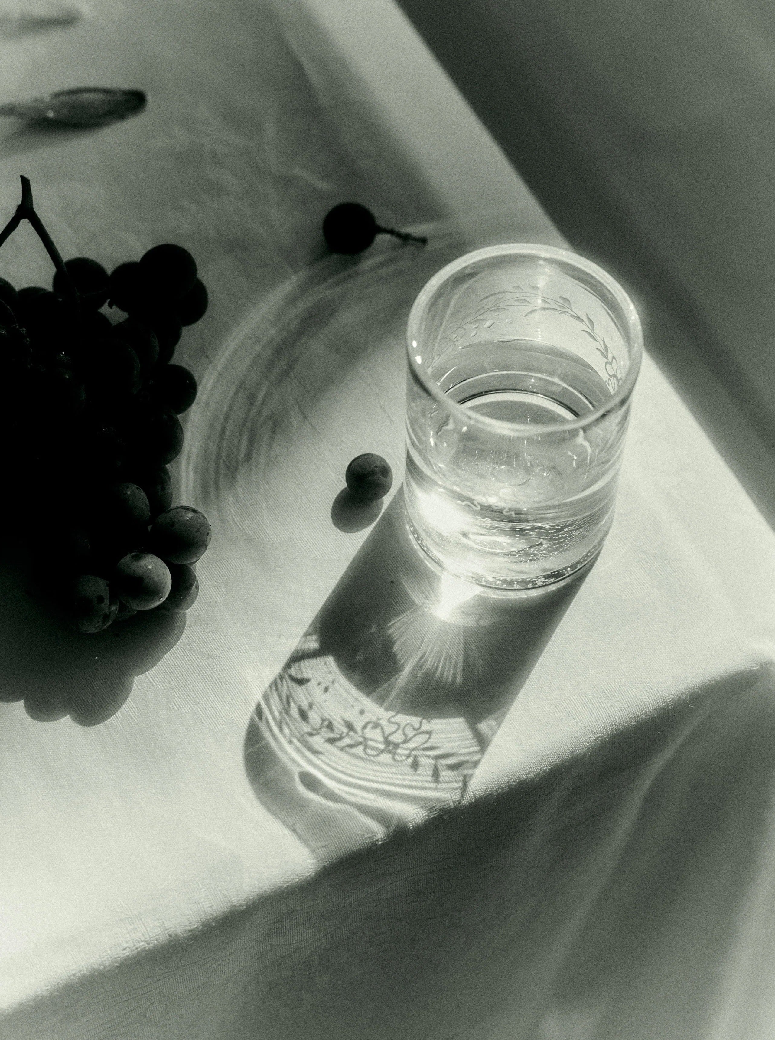 A monochrome image capturing the serene interaction of light and shadows on a table with a glass of Akua Objects Barbro Water Glass - Set of four, creating a reflection, accompanied by grapes nearby.