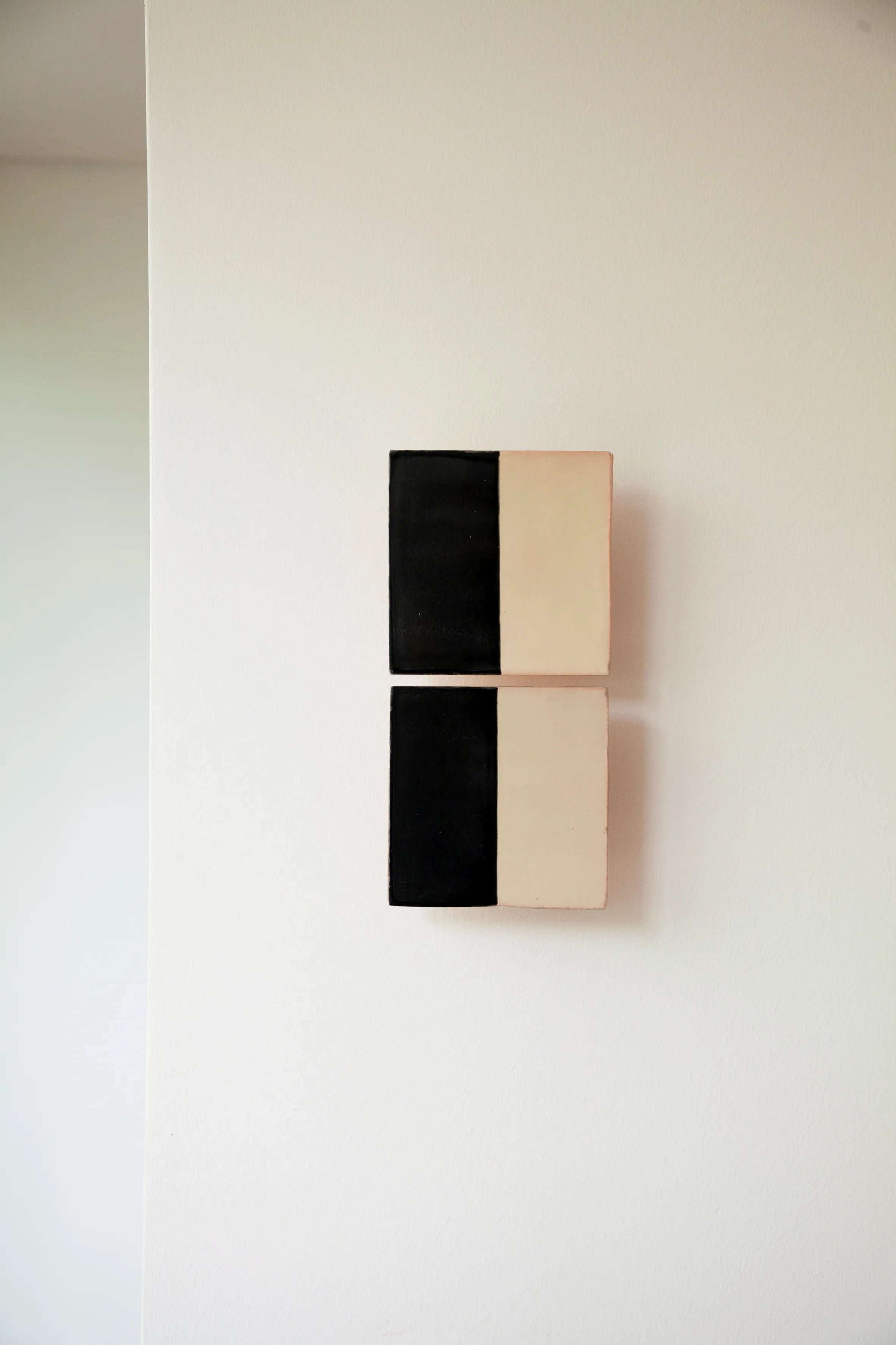 Two small, rectangular Tiles Line N pieces by Violaine d'Harcourt hang vertically on a white wall, featuring a black half on the left and cream on the right, capturing the minimalist symmetry typical of handmade artisanal creations.