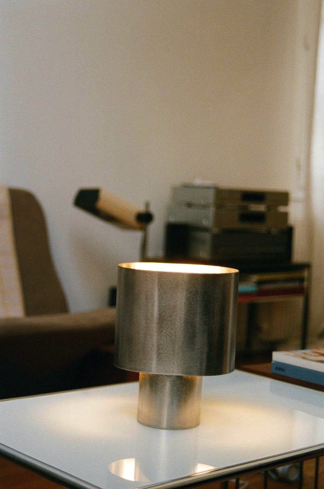 The Bien Paris Table Lamp in Brushed Steel sits on a white surface in a cozy, softly lit room with a brown chair, books, and audio equipment blurred in the background, highlighting its timeless design.