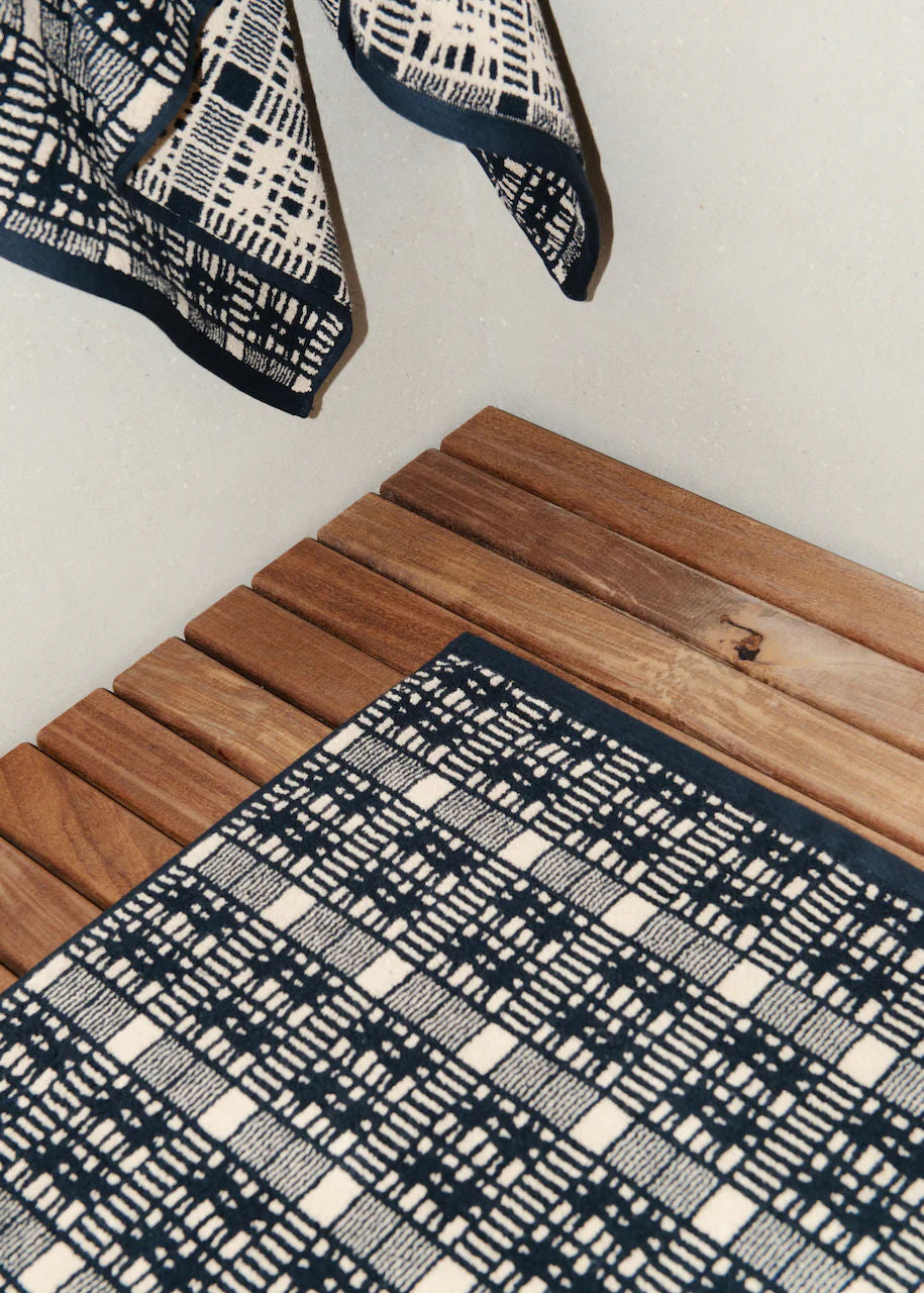 A checkered blue and white fabric Alma Bath Mat partially draped over a textured wooden surface, with part of the scarf suspended in the air.