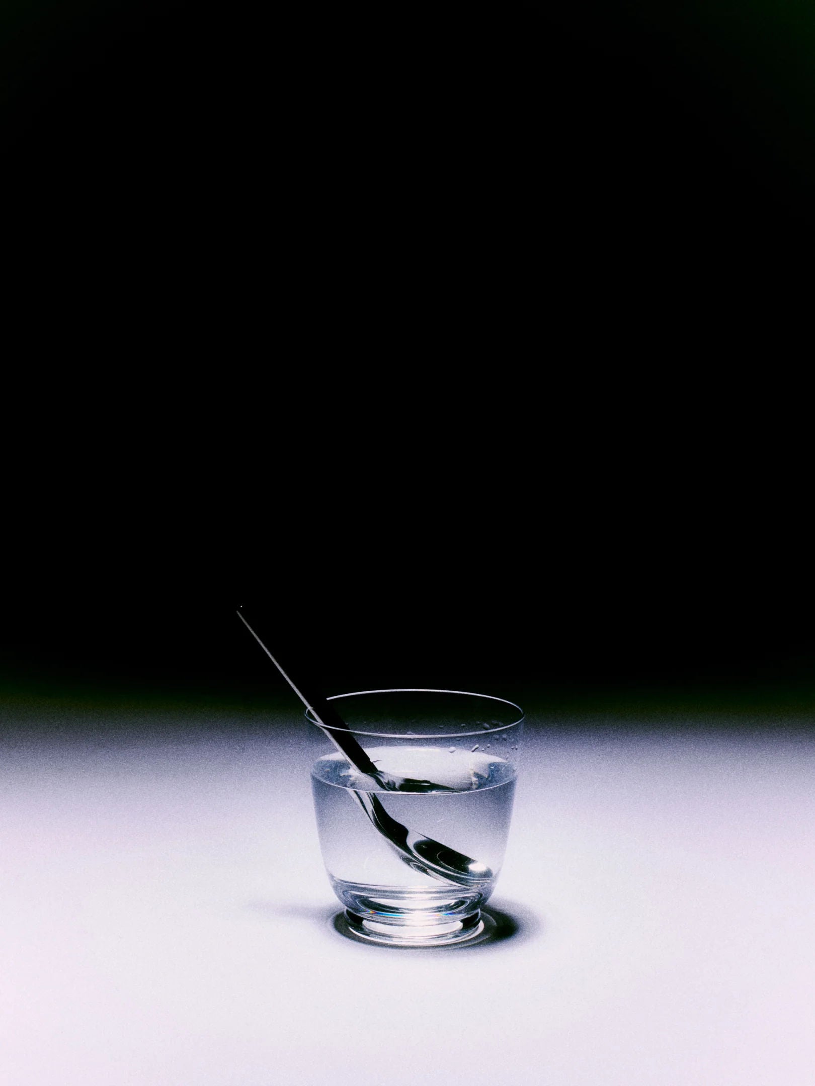 A glass of Walter 01 lead-free crystal water from Service Projects, with a silver spoon inside, resting on a white surface against a dark background. The spoon is partially submerged, creating a refracted image within the water, reminiscent of Art Deco elegance.