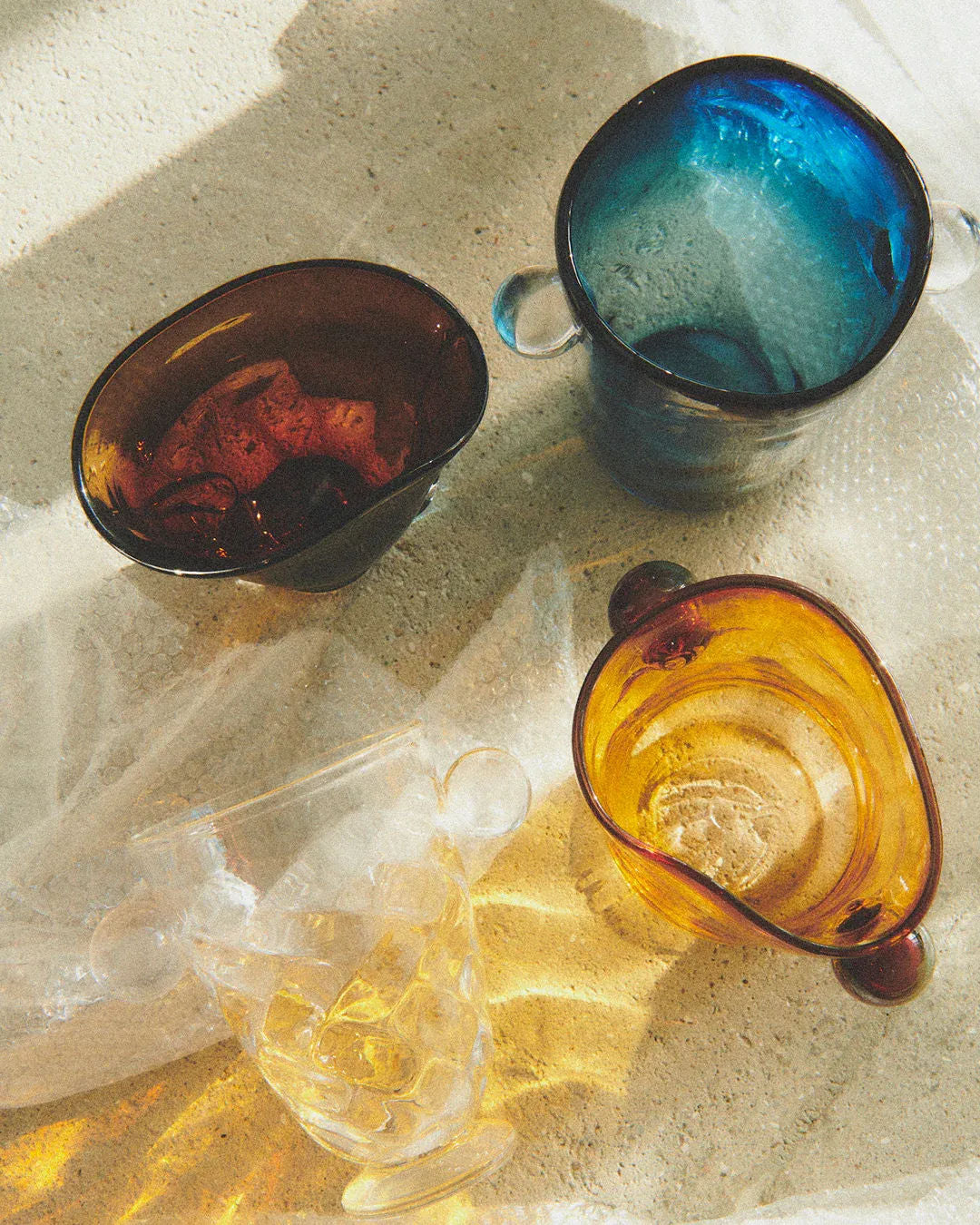 Four Karl Vases from Akua Objects in brown, blue, clear, and amber sit on textured concrete. Sunlight casts shadows highlighting their irregular shapes and thick handles. Reminiscent of Bohemian Glassware craftsmanship, bubble wrap peeks out underneath.