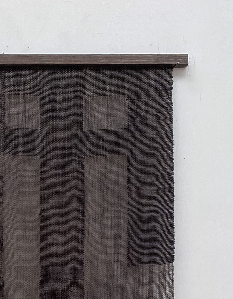 The "Grid I" tapestry by Sundance Studio hangs vertically on a white wall. Reflecting Bauhaus style, it showcases geometric patterns with intersecting lines and rectangles in a grid-like design, attached to a wooden rod.