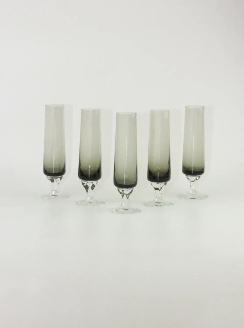 Five champagne flutes with gradient black-to-clear glass design and smoky gray stems, arranged in a row against a plain white background. The distinct tapering shape and elegant base highlight their modern aesthetic. This is the Set of Five Vintage Flute by Boga Avante Shop.