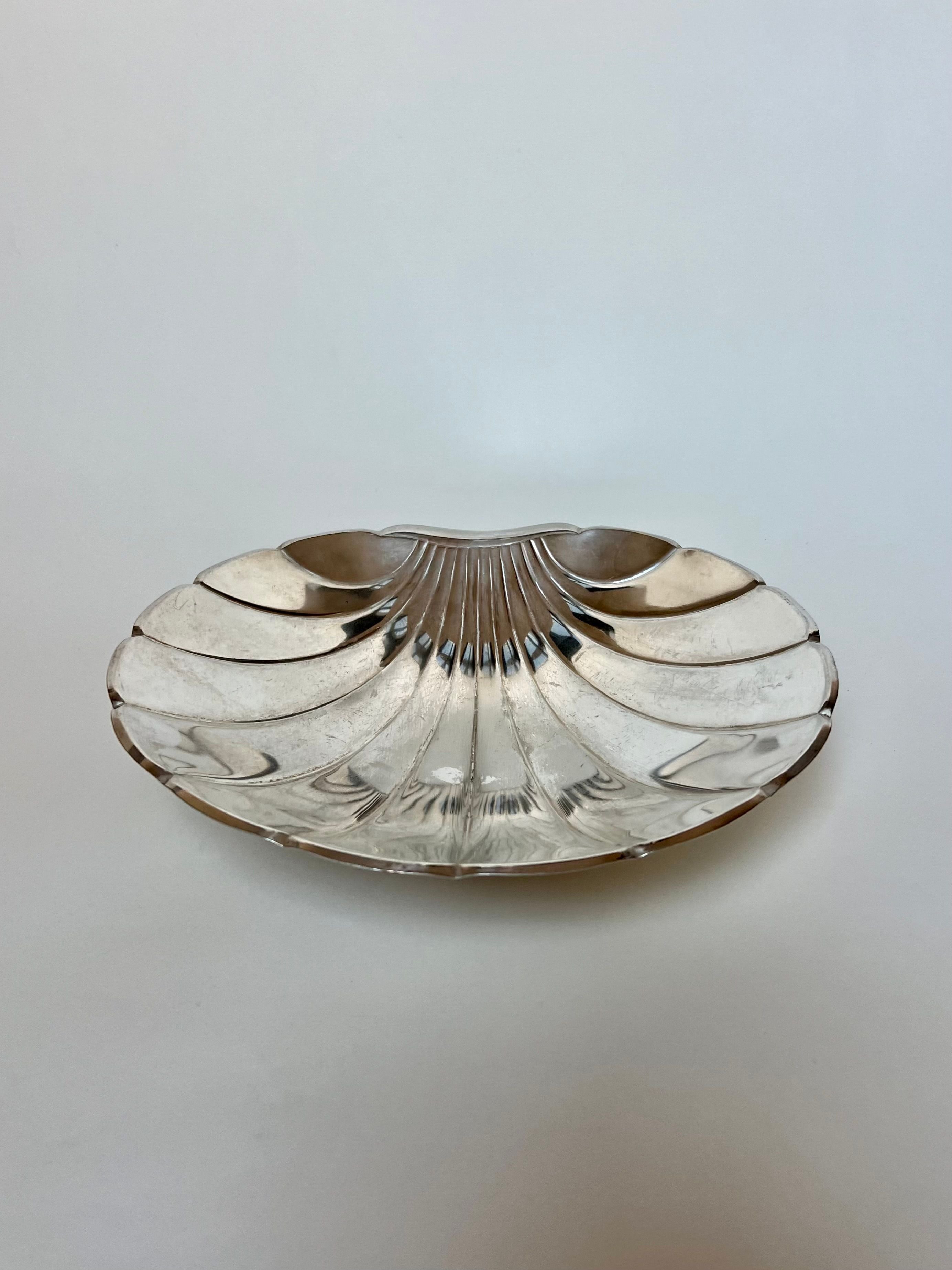 The Grand Silver Shell Dish by Manushi, with its shiny, silver-colored clam shape, ribbed texture, and ball feet, elegantly reflects light against a plain white background. This sophisticated design is sourced from France.