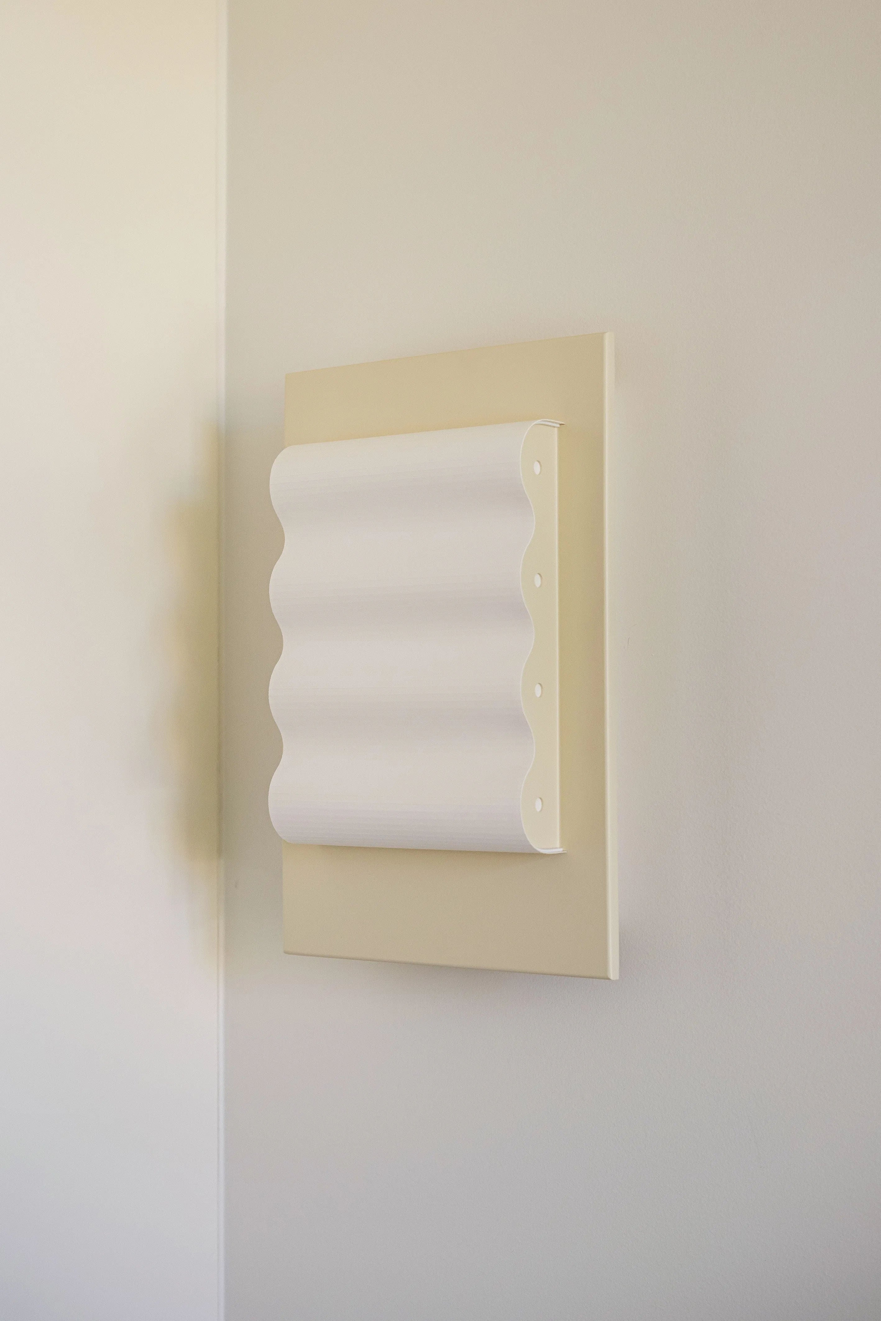 The Wall Light Frame OW S by Violaine d'Harcourt is a handcrafted, minimalist wall-mounted fixture with a wavy, sculptural design. Its vertically pleated pattern on a beige rectangular base contrasts beautifully against light-colored walls, serving as an elegant artistic sconce.
