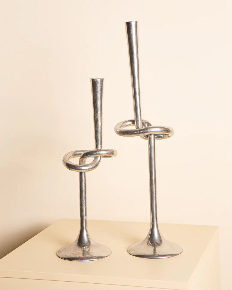 Two silver 80s aluminum "Nœud" candleholders by Treaptyque, featuring unique twisted designs, are displayed side by side on a light surface against a beige background. These sculptural pieces vary in height, with the taller one having a more prominent twist.