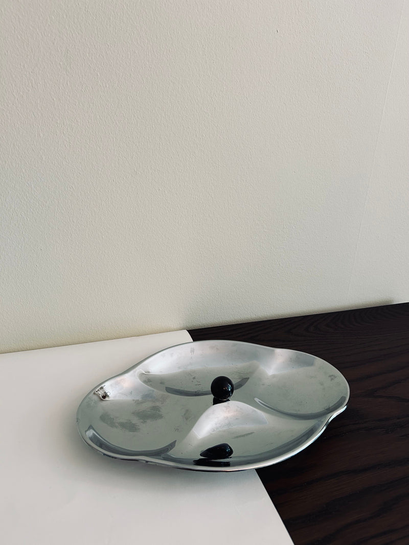 A Mid Century Serving Tray by Flock Studio, featuring curved sections and two small black objects, sits elegantly on a surface with white and dark wood patterns against a plain light wall.