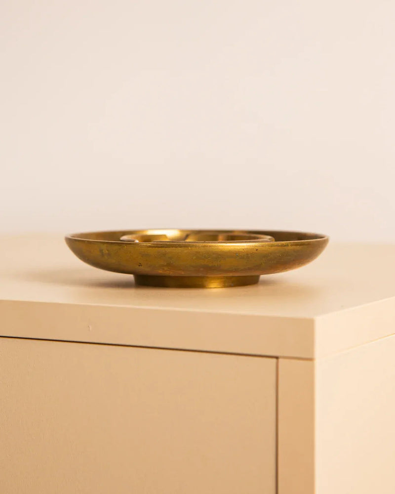 The Treaptyque Italian Circular Brass Ashtray 70's is elegantly displayed on a beige wooden surface, showcasing its high-quality Italian craftsmanship. This piece, with its flat, circular design, includes a central support for incense sticks set against a soft, neutral-toned background.