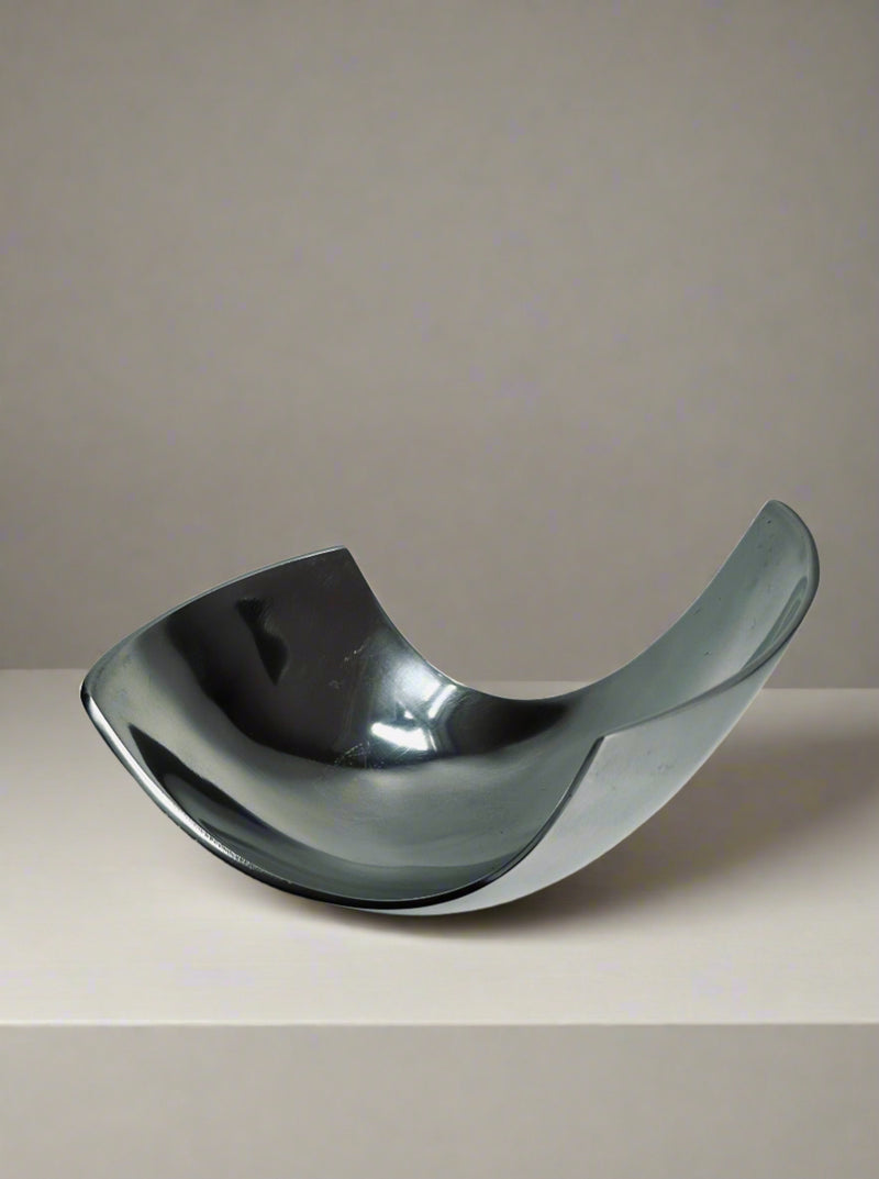 The Modernist Decorative Dish by Flock Studio features a sleek, reflective aluminum design with a distinctive curved shape, elegantly displayed on a plain surface against a neutral background. Its upward-curving sides create a modern, abstract look that is perfect for showcasing fruit.