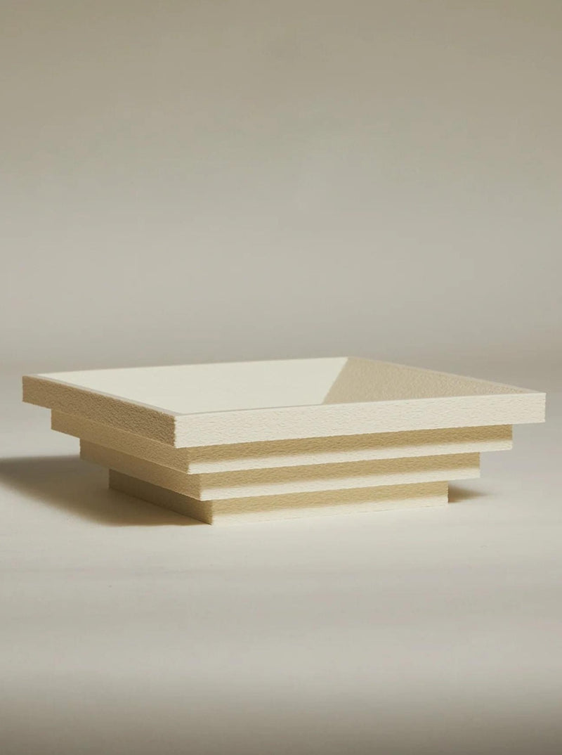 The Bau Bowl by Argot Studio, a sculptural cream-colored multi-tiered bowl with a geometric design, rests on a neutral surface. Its layers create a stepped effect for a modern, architectural look. Handcrafted to perfection, the softly lit and blurred background enhances its minimalist aesthetic.
