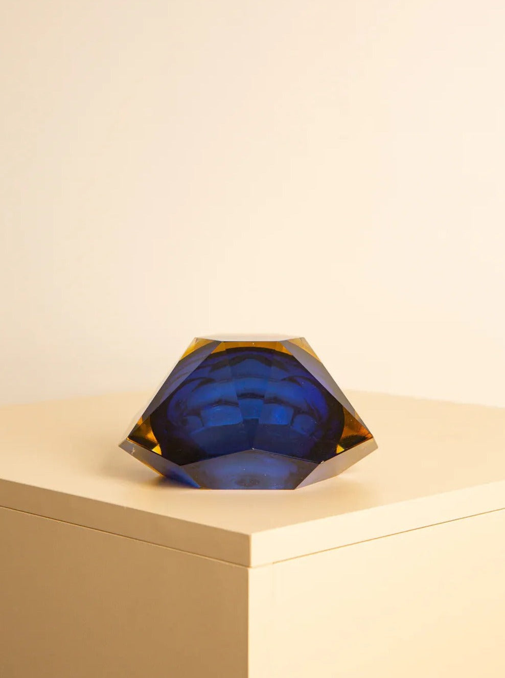 A Large vide-poches blue "Diamond" by Flavio Poli 60's from Treaptyque, handcrafted from fine Italian Murano glass, sits atop a beige pedestal. The sculpture has a faceted design and appears to be multi-dimensional, reflecting light in different shades. The background is a plain, neutral-colored wall.