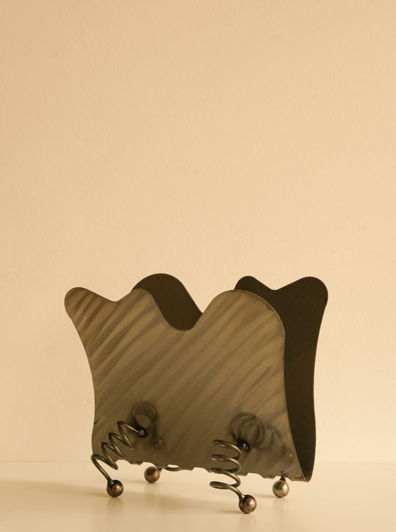 An artistic sculpture resembling a Les Objets Amsterdam Wavy magazine rack crafted from heavy metal, supported by small spherical feet, displayed against a plain light background with soft shadows.