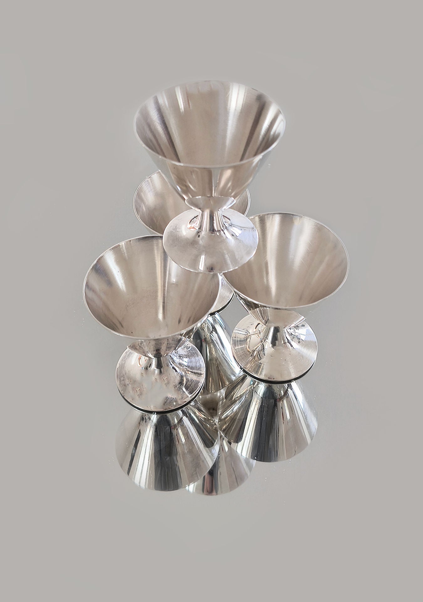 A set of six luxurious, silver-plated Christofle cups by Les Objoies is displayed on a reflective surface, casting mirrored reflections. The light gray background enhances their sleek and shiny texture, making them an exquisite addition to any home decor.