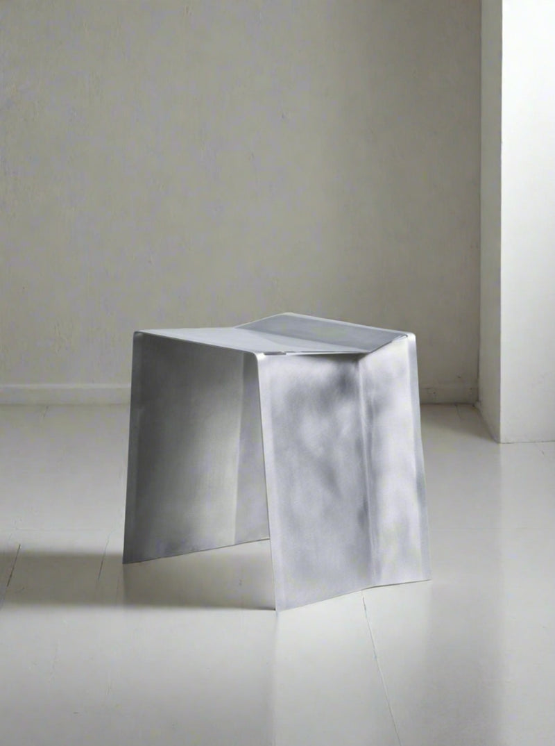 A minimalist Paul Coenen Camber Stool, featuring angular cuts and an inverted trapezoid shape, sits on a concrete floor against a plain gray background.
