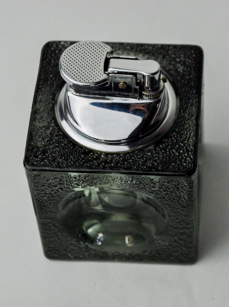 The elegant Ice Cube Lighter by Antonio Imperatore from Malamar Studio features a vintage metal top with a flip cover and circular ignition area, complemented by a textured, square black Murano glass base. Resting on a light, plain surface, this sophisticated lighter exudes the timeless charm of Antonio Imperatore's designs.