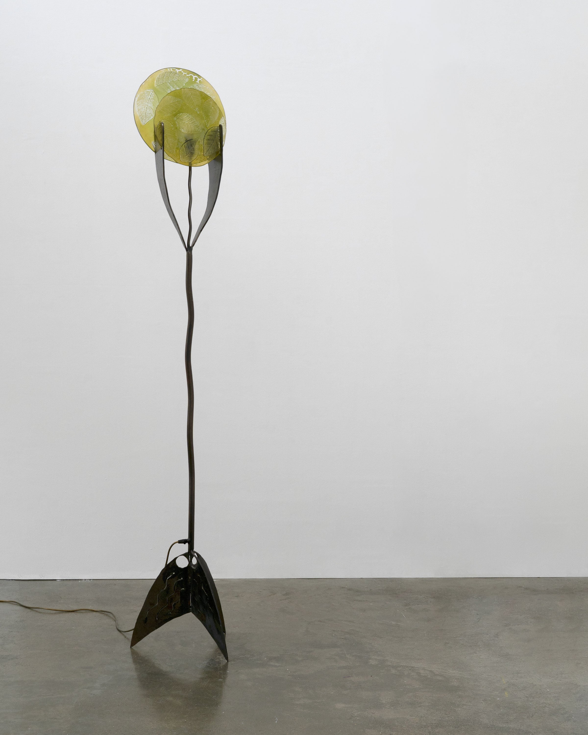 The Brutalist Wrought Iron and Cast Glass Lamp by Tom Bogle, Norway, c.1970, features a wavy black stand, triangular wrought iron base, and round yellow-green shade on a gray concrete floor against a white wall.