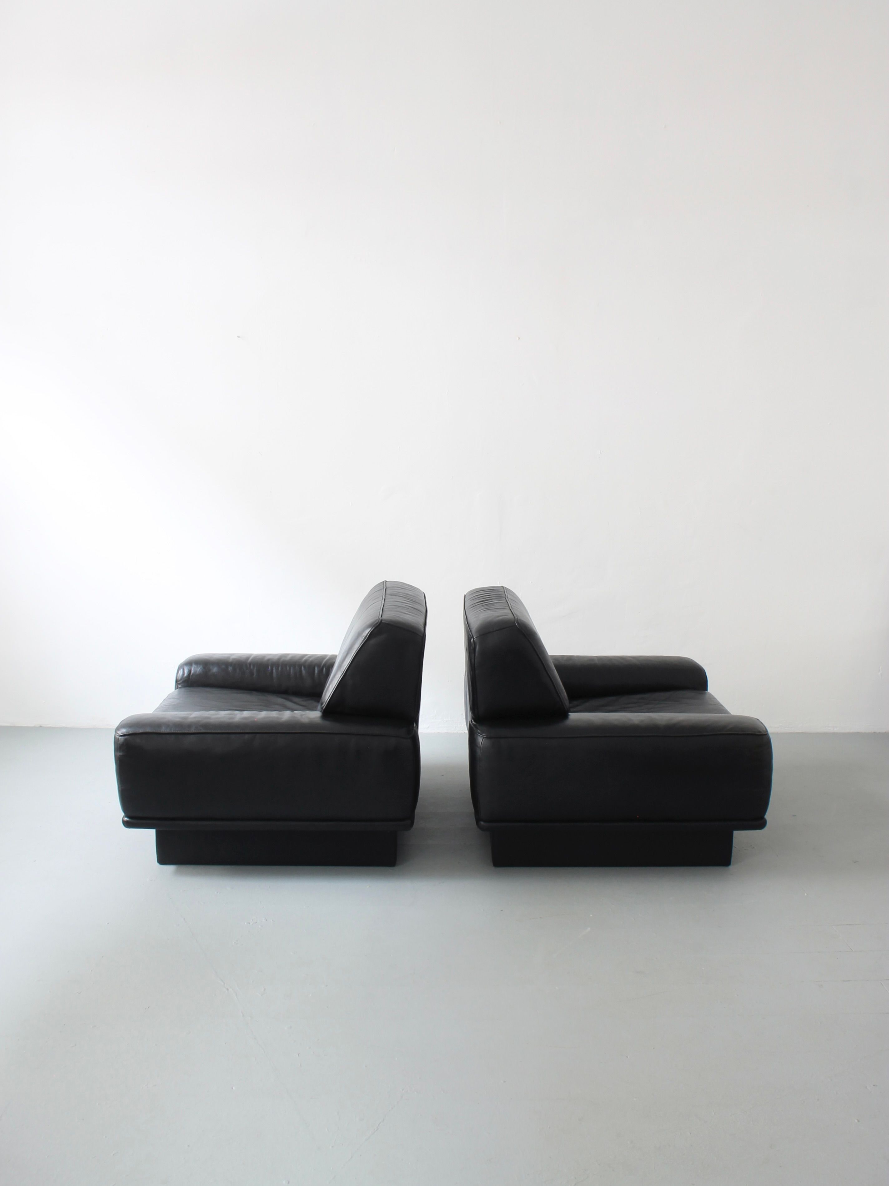 Two black leather lounge chairs by Afterhour Furniture, crafted from high-end leather, are set back-to-back against a plain white wall on a light gray floor. The image features Swiss luxury furniture with a minimalist, modern design known for its clean lines and simple geometry.