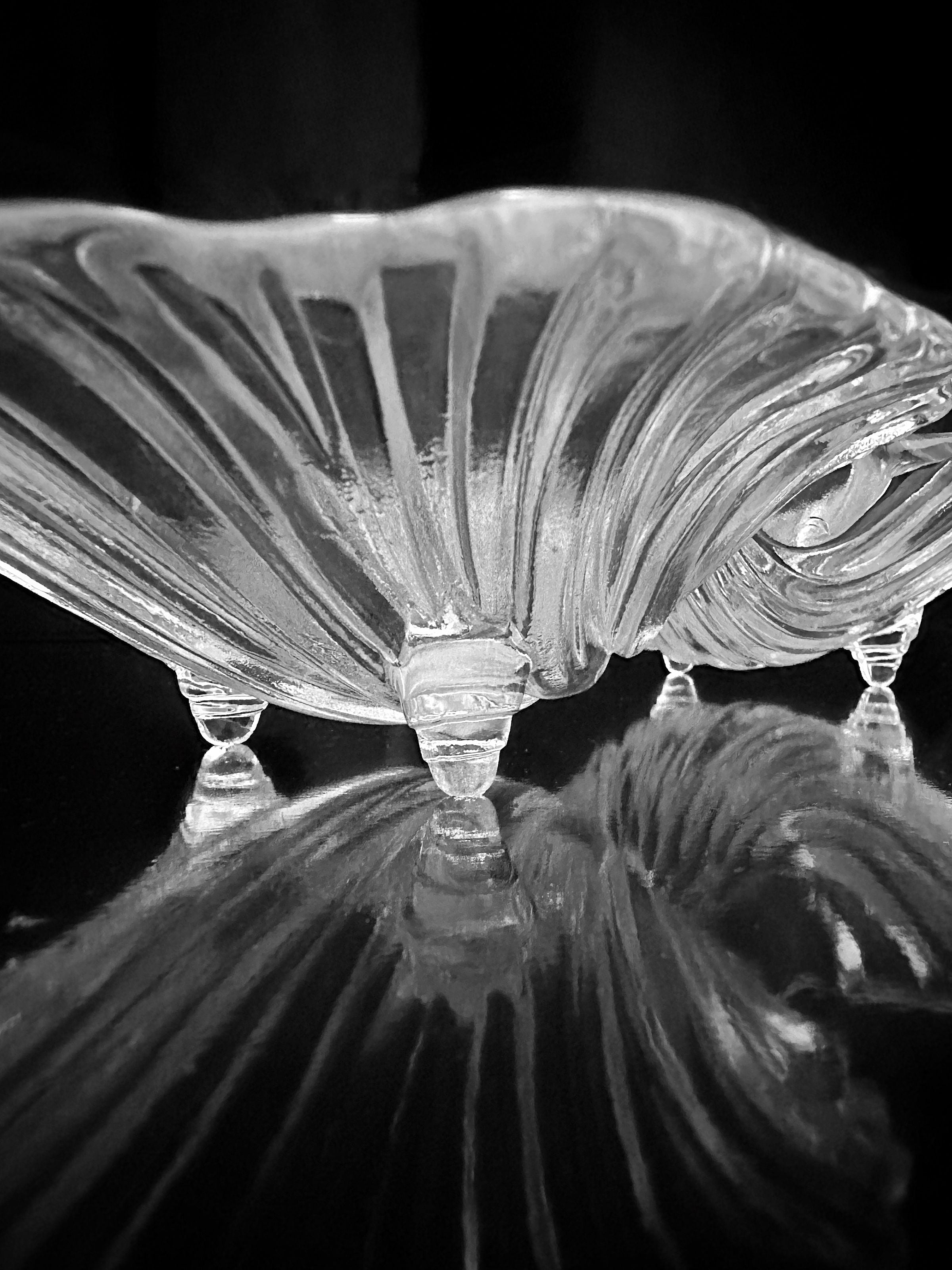 A black and white image showcases an elegant, ornately designed glass dish from the Dodo Vintage 1980s Shell Shaped Glass Bowls collection. The dish features four small feet and casts a detailed reflection on a shiny surface. With its wavy, shell-like pattern, it's perfect for presenting artistic delicacies, offering a sophisticated appearance.