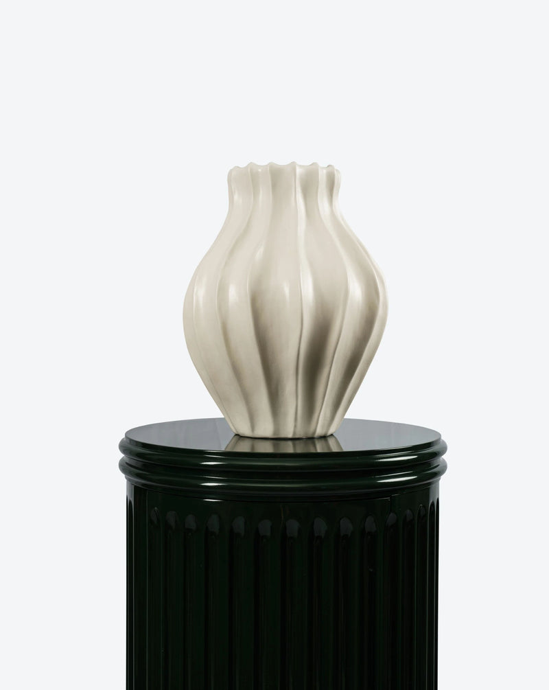 A Medium Cloak Vase by Ransom & Dunn, featuring a sculpted wavy design in white, is displayed atop a glossy black cylindrical pedestal against a light background. This exquisite piece is part of an exceptional ceramic collection and is proudly handmade in Portugal.