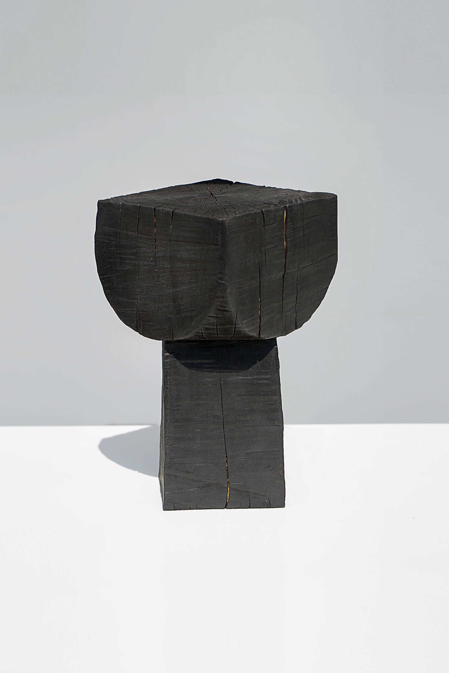 Touch With Eyes' Burnt Black Coffee Tables/Stools features a minimalist design with a textured black block on a rectangular base, emphasizing its stark geometric form and rough surface against a plain backdrop.