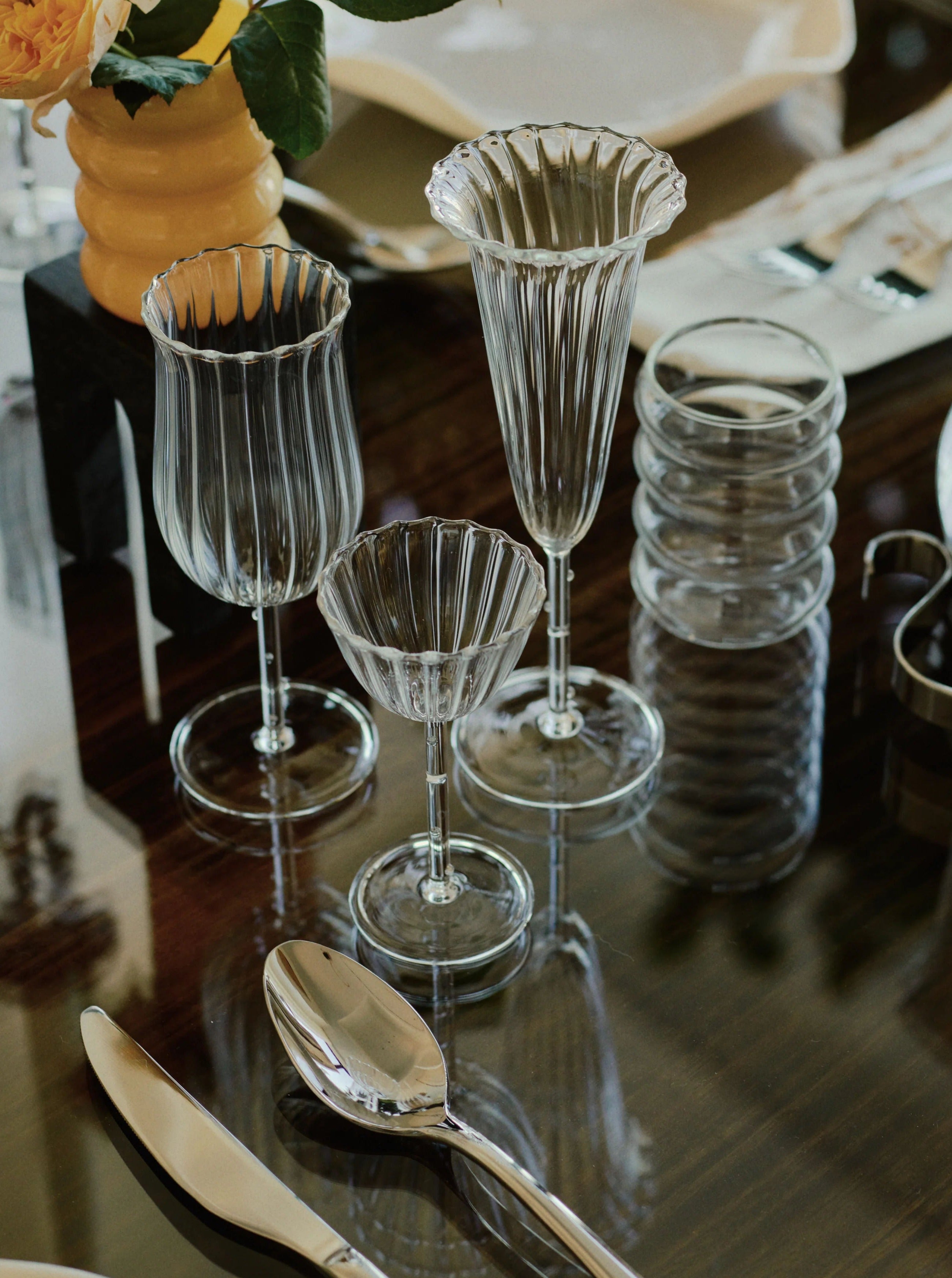 A close-up of a dining table set, featuring an array of elegant glassware, including Sophie Lou Jacobsen's Tulip Wine Glass - Set of Four that enhance the release of aromas, a cocktail glass, and stacked tumblers. The table also has polished silverware, a plate in the background, and a yellow vase with flowers.