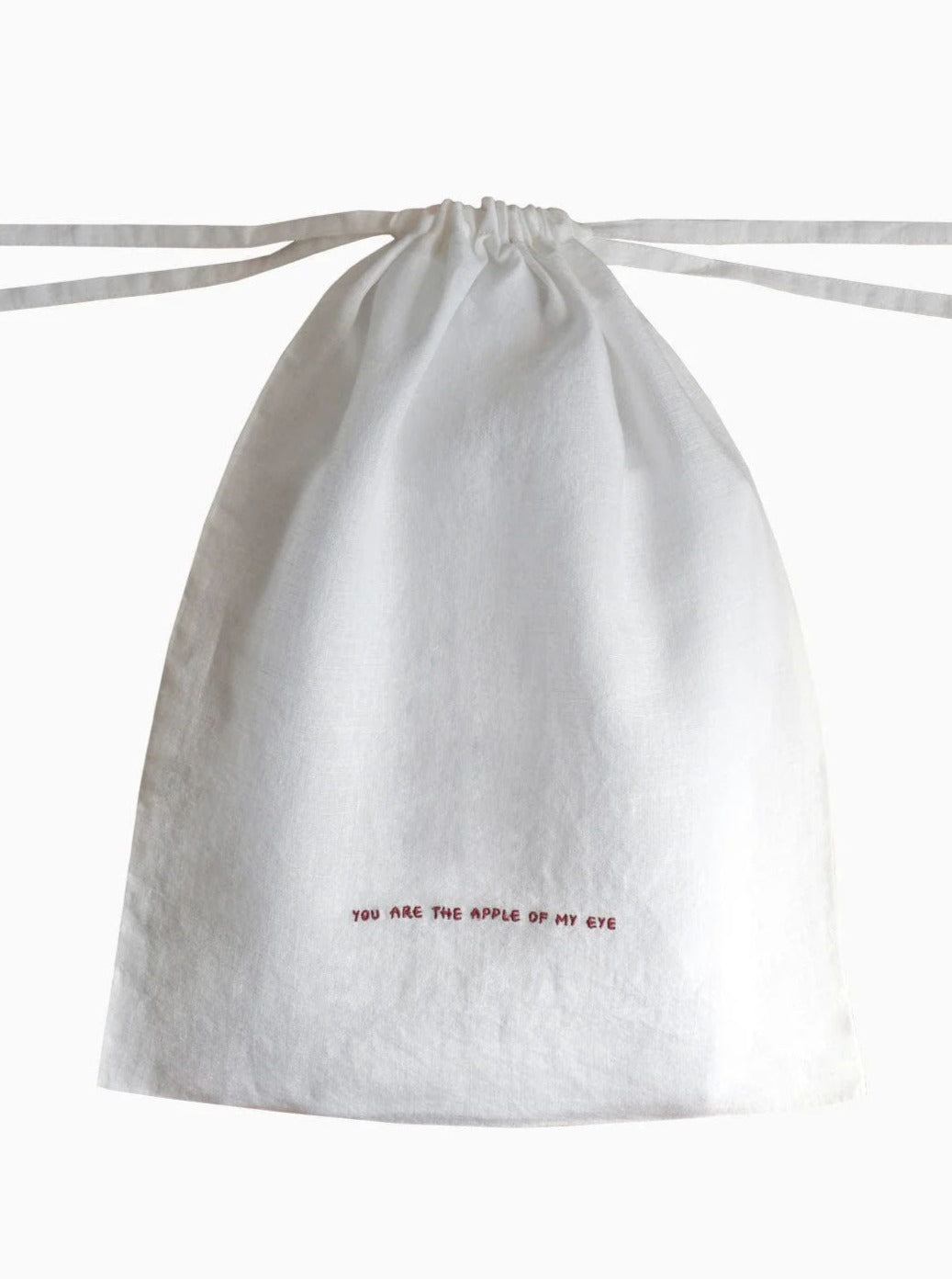 The Reusable Linen Produce Bag 'You Are The Apple Of My Eye' by arkitaip features a minimalistic design and is crafted from OEKO-TEX certified white linen. Embroidered near the bottom in small red letters is the phrase "YOU ARE THE APPLE OF MY EYE." The drawstrings are neatly tied at the top, making it perfect for keeping bread fresh.