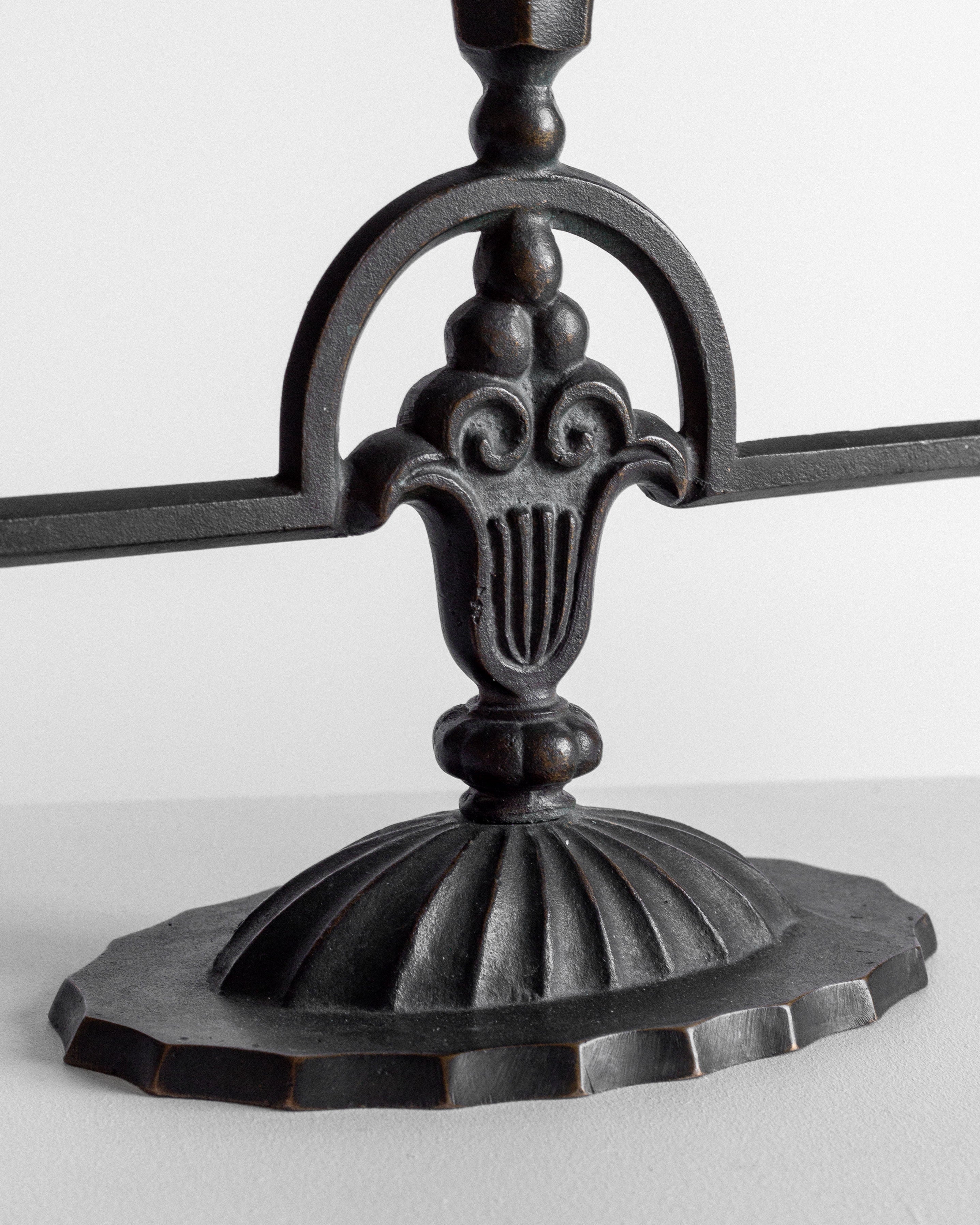 A close-up of the Spigel Art Deco Bronze Candelabras from the 1930s showcases an ornate black metal base, reminiscent of Swedish craftsmanship. The intricate detailing and symmetrical patterns are highlighted on the solid bronze base, which rests gracefully upon a textured, scalloped pedestal. This decorative piece beautifully blends curved and linear elements against a plain light gray background.