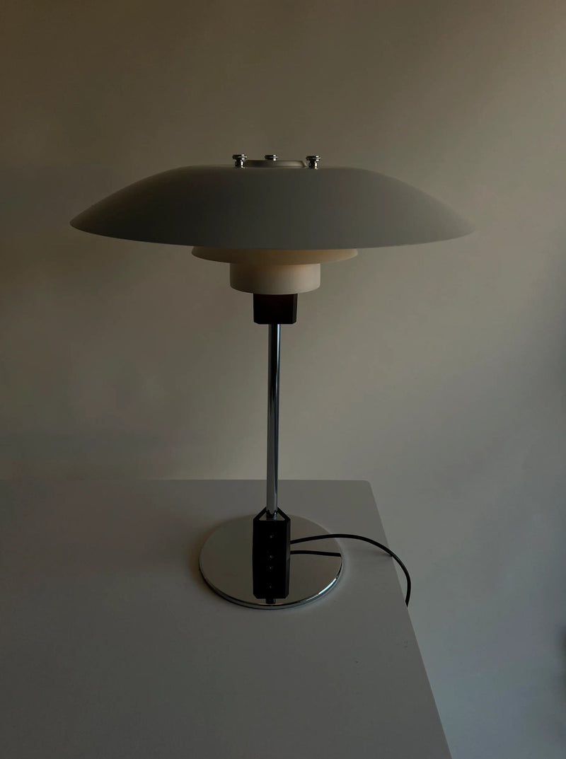 A sleek, modern table lamp with a wide, dome-shaped shade and a minimalist design stands on a round, chrome base. Reminiscent of Poul Henningsen's 1983 vintage table lamp designs, the CU-RA-TED Poulsen Table Lamp PH 4/3 1983 is placed on a plain, light-colored surface with a power cord extending from the base. Neutral background with soft lighting.