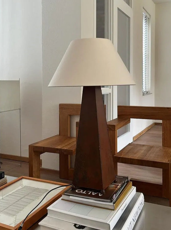 A minimalist, modern interior showcasing a wooden chair and table. On the table are stacked books and a CU-RA-TED Obelisk Table Lamp with a white shade. The background has white walls and a window with blinds, enhancing the airy and clean ambiance with refined sophistication.