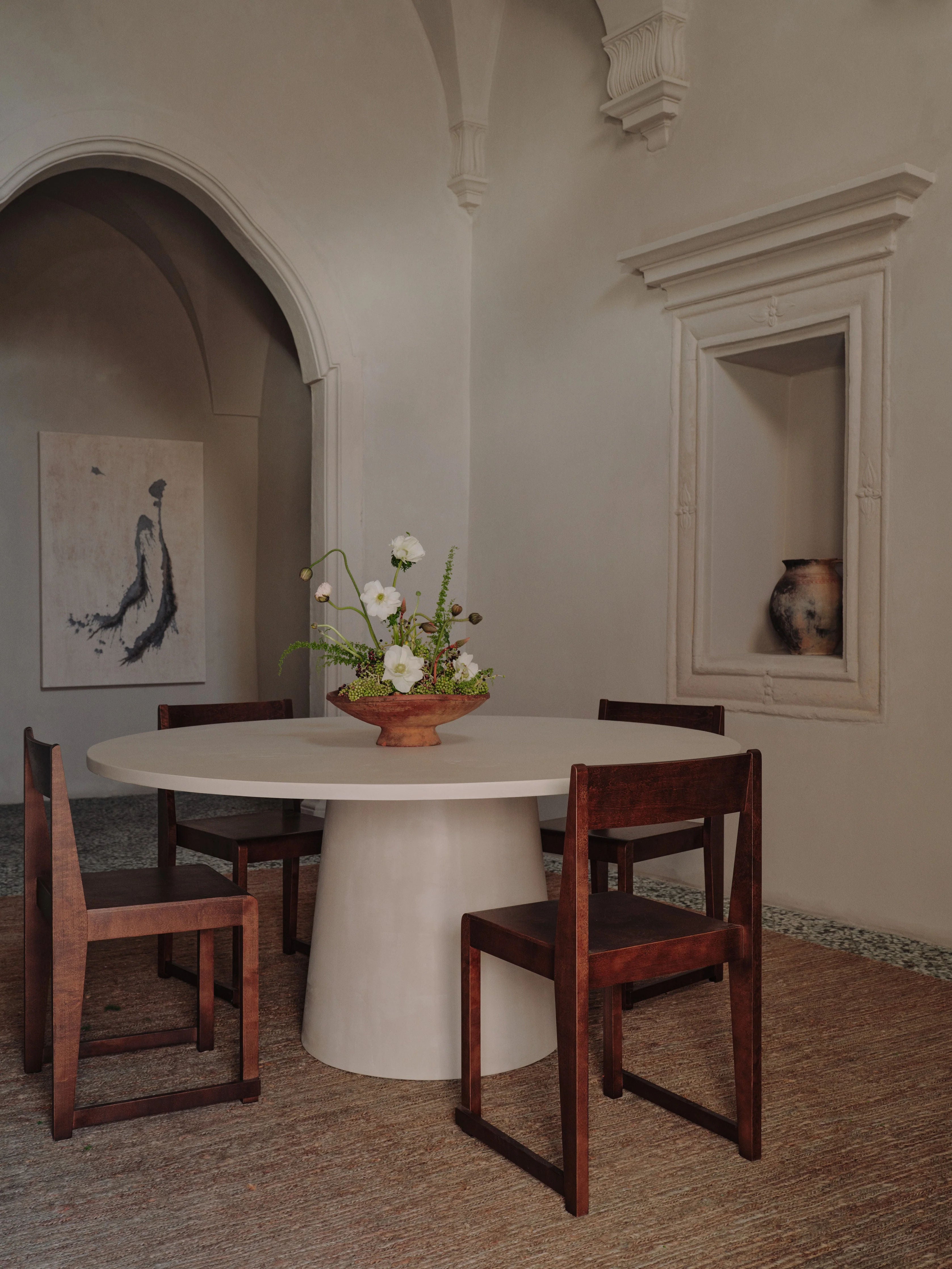 A minimalist dining room with a modern design features the MarlotBaus "Core" Dining Table, known for its scratch-resistant microcement top, surrounded by four wooden chairs and accented with a simple floral centerpiece. The room includes an arched doorway, a painting on the wall, and a niche showcasing a ceramic vase.