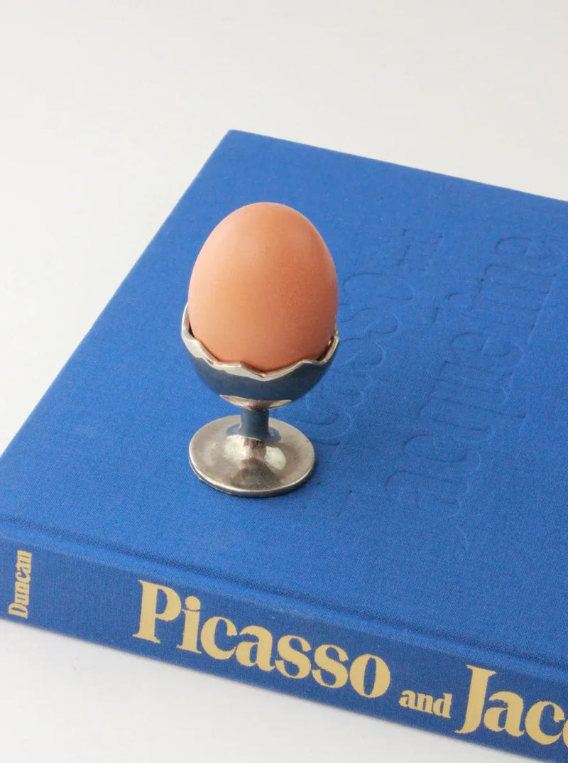 A brown egg sits in an Egg Cup by Boga Avante Shop placed on top of a blue hardcover book titled "Picasso and Jacqueline." The egg and egg cup are centered on the book, which lies on a white surface. The silver cuff of the Egg Cup by Boga Avante Shop features a decorative edge that adds a touch of elegance.