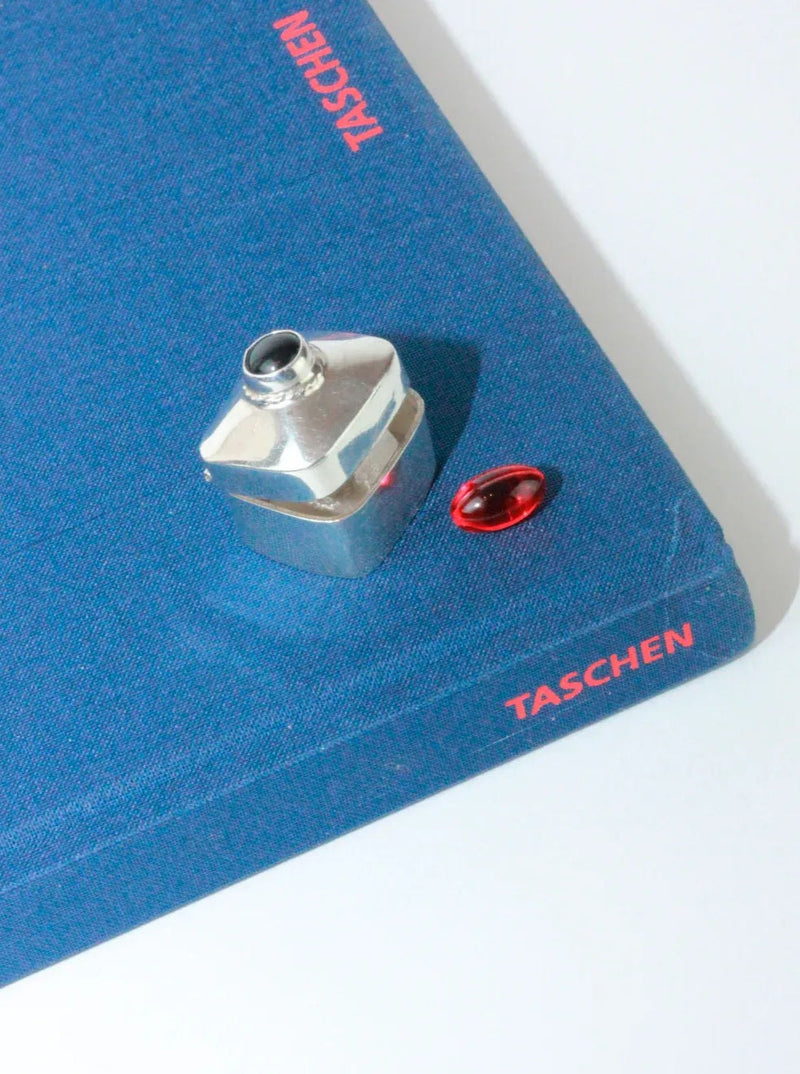 A close-up of a corner of a blue book with 'TASCHEN' written in red on both the spine and cover. On the book, there is a small handmade Taxco Mexico metallic tank-shaped object and Boga Avante Shop's Silver Pillbox With Onyx Stone placed next to each other. The background is a plain white surface.