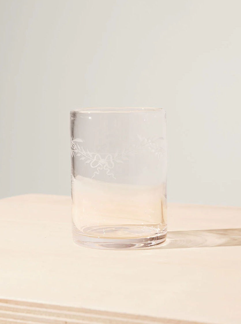 A transparent Akua Objects Barbro Water Glass Large half-filled with water, positioned on a light beige surface against a plain pale background. The glass has a delicate floral etching on its side.