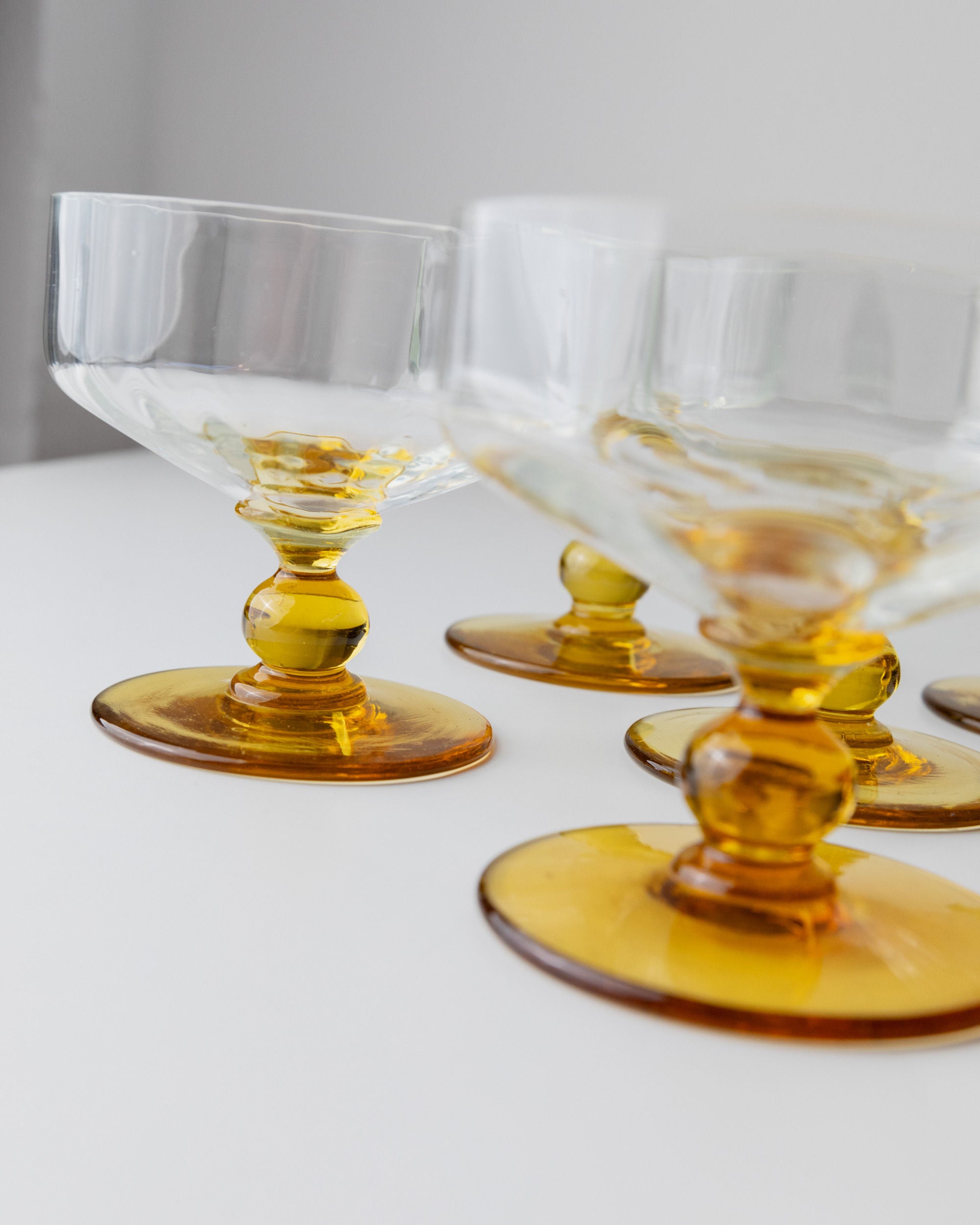 A set of 6 Murano glass goblets from Bottega Jacobs, featuring clear bowls and amber stems from the 70s Italy collection, are placed on a white surface. The light catches their intricate design, highlighting the elegance in a stylish display.