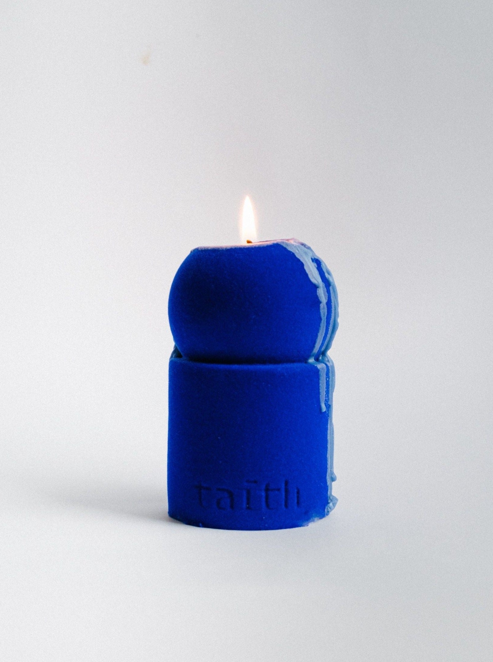 A lit Lulu Electric Blue Velvet Edition soy candle with a unique design appears against a plain white background. Wax has begun to melt and drip slightly down the side, showcasing the meticulous detail. The brand name "Taith" is embossed at the base, highlighting its Velvet edition quality.