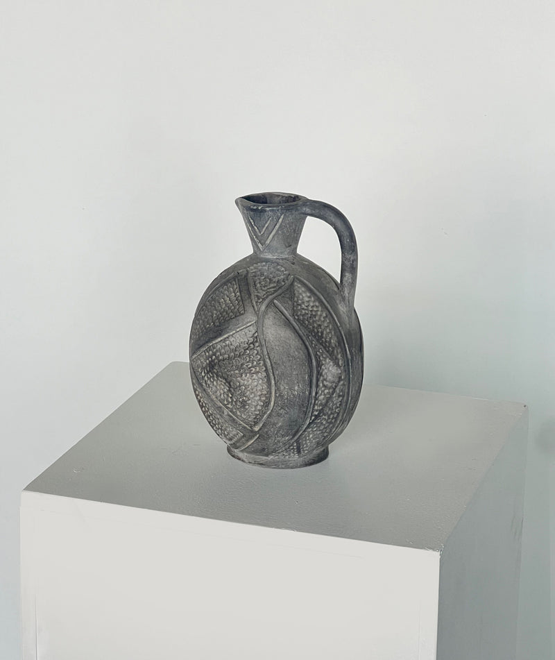 A handcrafted round ceramic jug from Porter Studio, named "Jug with Hand Markings," sits on a white pedestal against a plain background. This intricately carved piece features a handle and spout, adorned with abstract grey patterns that give it a vintage, artistic appearance reminiscent of artisan pottery.