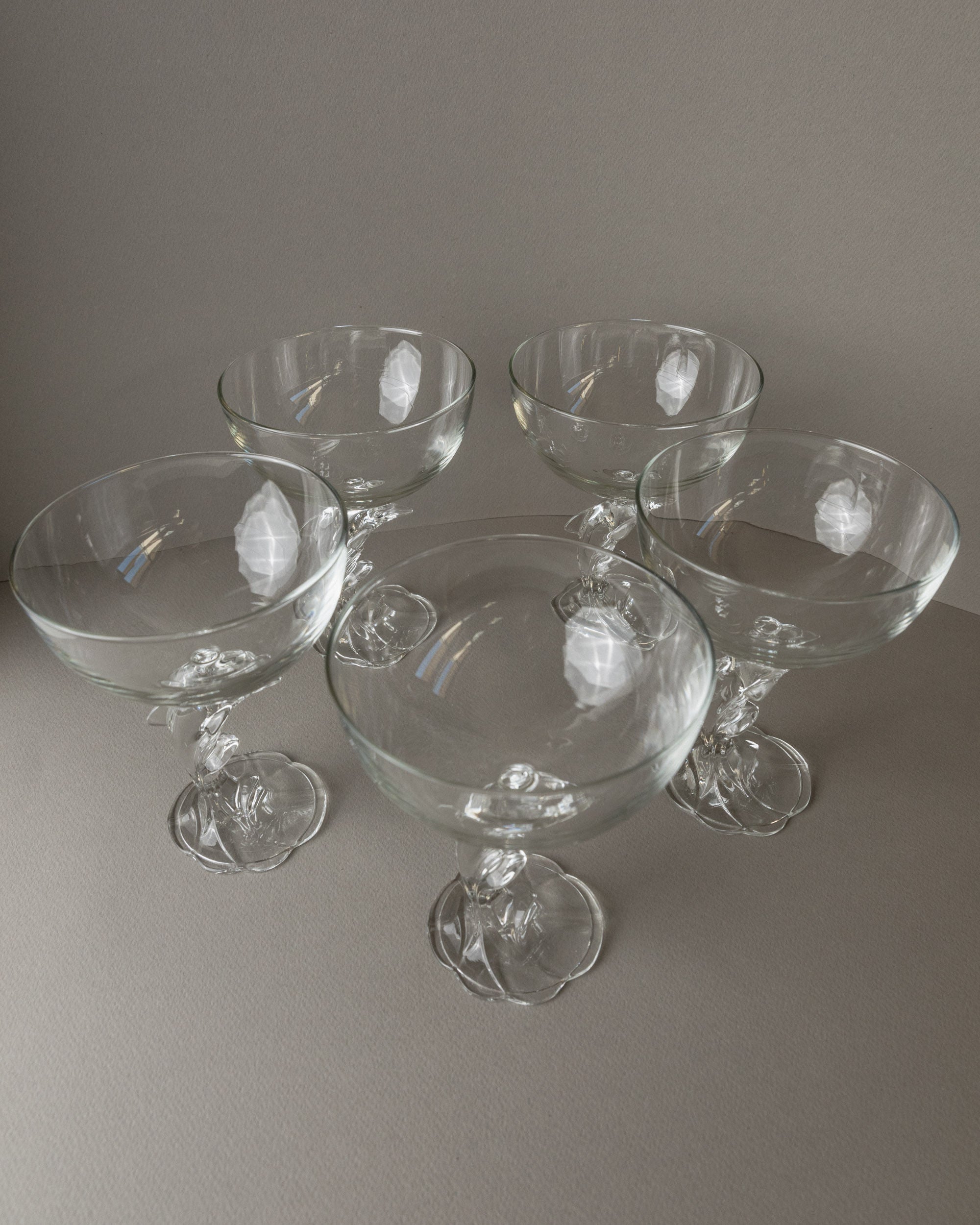 Five Luminarc Delfin Champagne Coupes by Bottega Jacobs, crafted in 80s France, feature ornate stems and are arranged on a neutral-toned surface. Their subtle shadows and a plain background highlight the elegance of these vintage glasses.