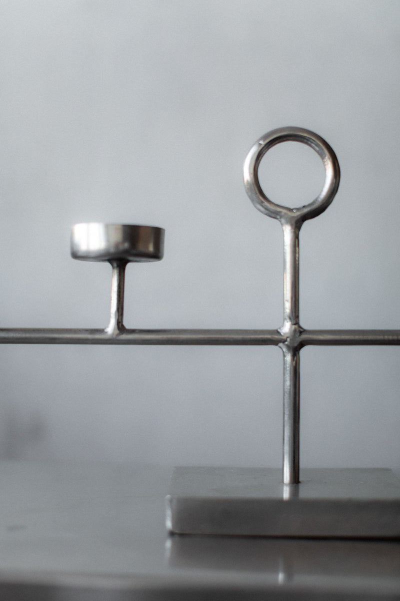 The Out For Lunch Steel Candleholder is a minimalist metal sculpture with a modern design, featuring two vertical elements: one has a small circular platform ideal for holding candles, while the other forms a round loop. It sits on a flat base against a neutral background.